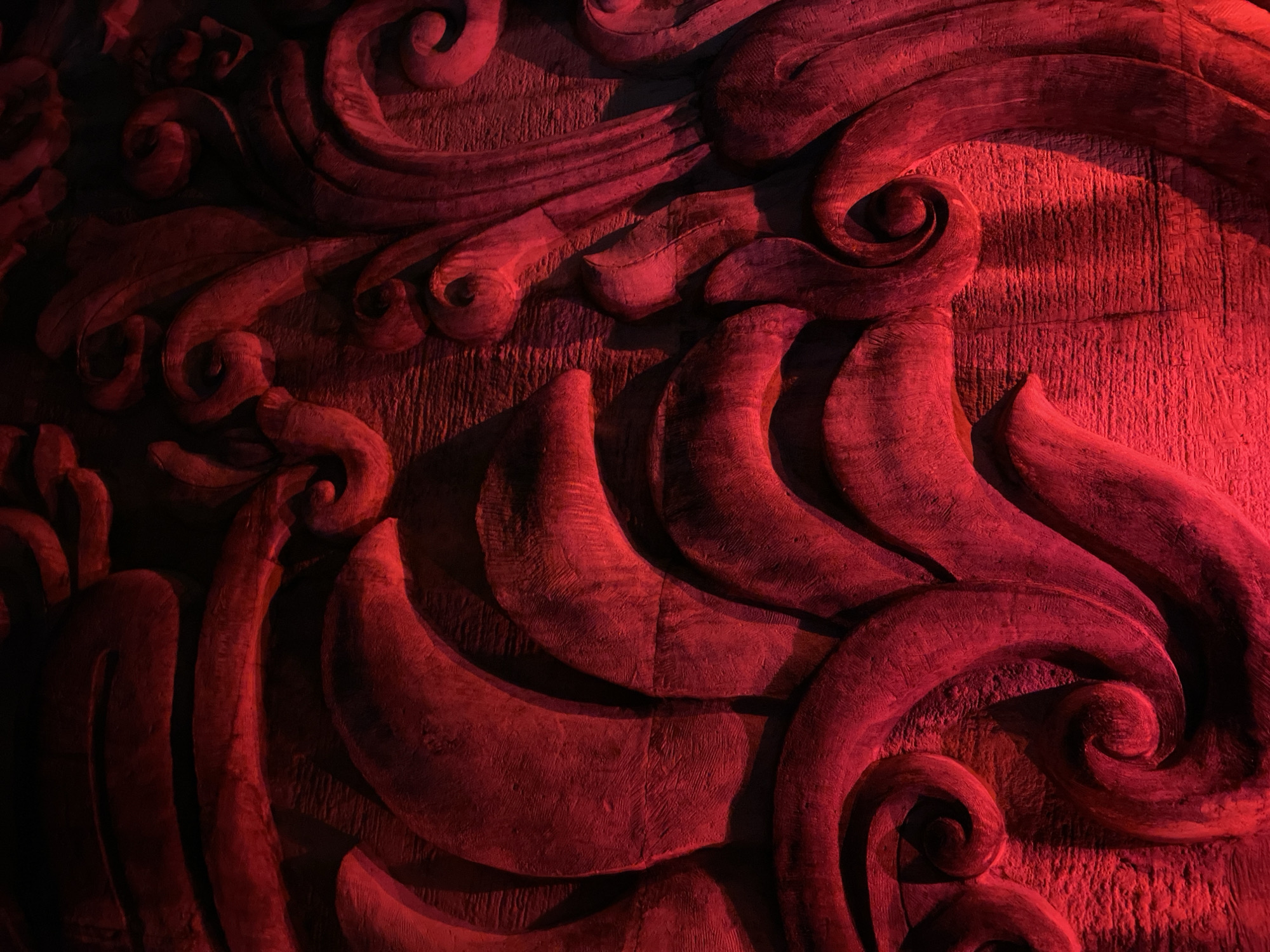 A close-up, abstract photograph captures a section of a textured, carved surface bathed in a dramatic red light. The carving features organic, flowing shapes reminiscent of stylized leaves, petals, or waves, with prominent curves and swirls. The texture of the underlying material, possibly stone or wood, is visible as a rough, linear pattern. The lighting creates deep shadows and bright highlights, accentuating the three-dimensionality of the carving and giving it a somewhat ominous or theatrical appearance. No people, animals, or distinct objects are identifiable in the image. The environment is entirely defined by the carved surface and its illumination.