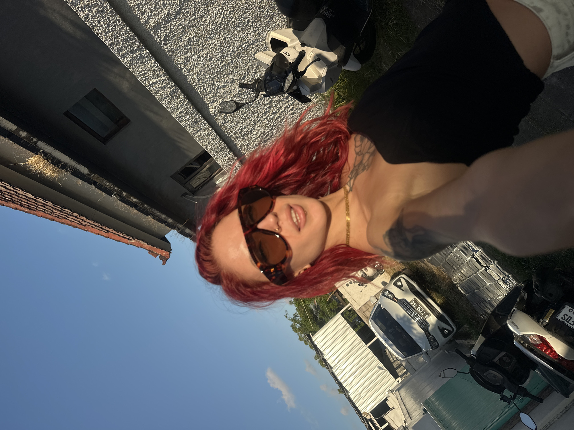 A woman with red hair and sunglasses takes a selfie. She is wearing a black top and a gold chain. She has tattoos on her neck and arm. Behind her is a building, a motorcycle, a car, and a blue sky. The license plate on the white car reads "B 2753 TRD". The license plate on the motorcycle is partially visible and reads "FDS 503". Based on the license plates, it is likely taken in the city of North Kuta, Indonesia.
