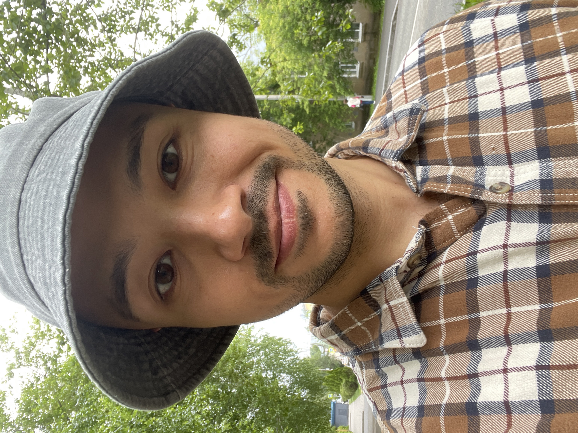 The image captures a male individual in an outdoor urban setting in Izhevsk, Russia, during daytime. The primary subject is a male with a medium complexion, dark brown eyes, and a short, dark beard. He is wearing a grey, textured fabric bucket hat and a brown, cream/off-white, dark blue, and reddish-brown plaid flannel shirt. He is positioned facing the camera, exhibiting a slight smile.
The background on the left side features lush green deciduous trees, a paved sidewalk, a dark blue rectangular utility box, and a conical evergreen shrub. A street is visible further back. The background on the right side also contains green deciduous trees, a light-colored building with multiple rectangular windows, and a paved road. In the distance on the right, another individual is observed walking away from the camera, dressed in a light-colored top and dark pants. A utility pole stands on the right side of the frame. The lighting suggests an overcast or partly cloudy sky.