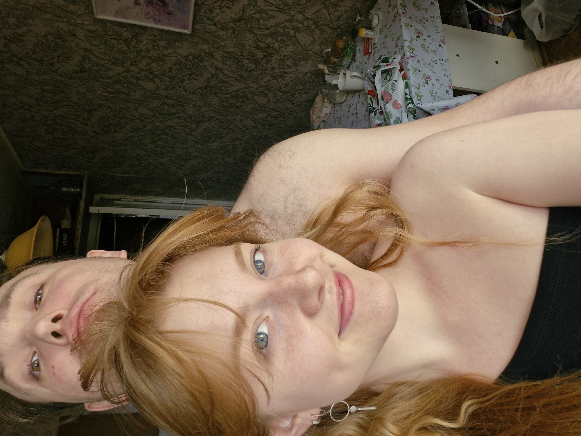 The image captures a candid, close-up moment between two individuals in an indoor setting, rotated 90 degrees counter-clockwise. In the foreground, a young woman with long, flowing red hair, fair skin, and noticeable freckles across her face looks directly at the camera with a soft, friendly smile. Her striking light blue eyes are prominent, and she wears a black top. A silver circular earring is visible in her right ear.
Behind her and partially to the left, a young man is seen from the chest up, with light brown hair and green or hazel eyes. He is looking away from the camera, appearing relaxed, and his right shoulder and upper arm, covered in light hair, are visible. A yellow object, possibly a hat, with the Cyrillic text "ЯРЛ" faintly inscribed, rests near his head.
The background on the upper side of the rotated image features dark, patterned wallpaper with a subtle tree or branch design, and a framed picture hangs on it. The background on the lower side shows a cluttered table or shelf covered with a white tablecloth adorned with a floral pattern. Various household items are visible, including white mugs, bottles, a clear bowl filled with fruit (likely oranges or lemons), and plastic bags, suggesting a lived-in home environment. The overall mood is intimate and casual.
