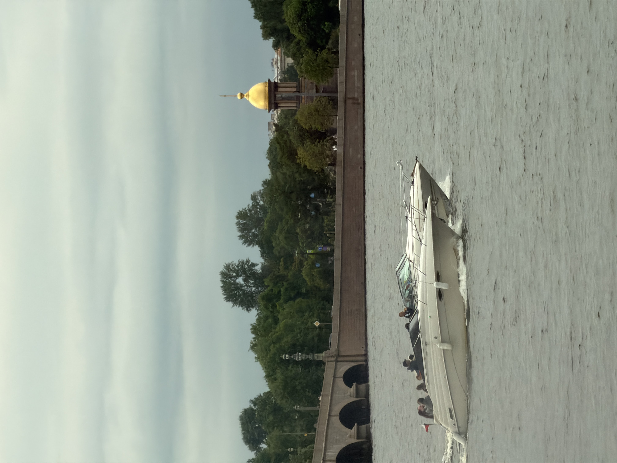 An outdoor, overcast, eye-level shot depicts two white motorboats on a body of water in front of a stone bridge and buildings in the background. The boat in the foreground is larger and closer to the viewer, with at least six people on board. Several individuals are standing or sitting, and one person appears to be steering. The second boat, slightly smaller and behind the first, also carries several passengers. The water is choppy, creating white wakes behind both vessels. The background features a long, arched stone bridge on the left, with trees and buildings lining the opposite bank. On the right side of the background, a prominent building with a golden dome rises above the trees. The sky is filled with gray, horizontal clouds.