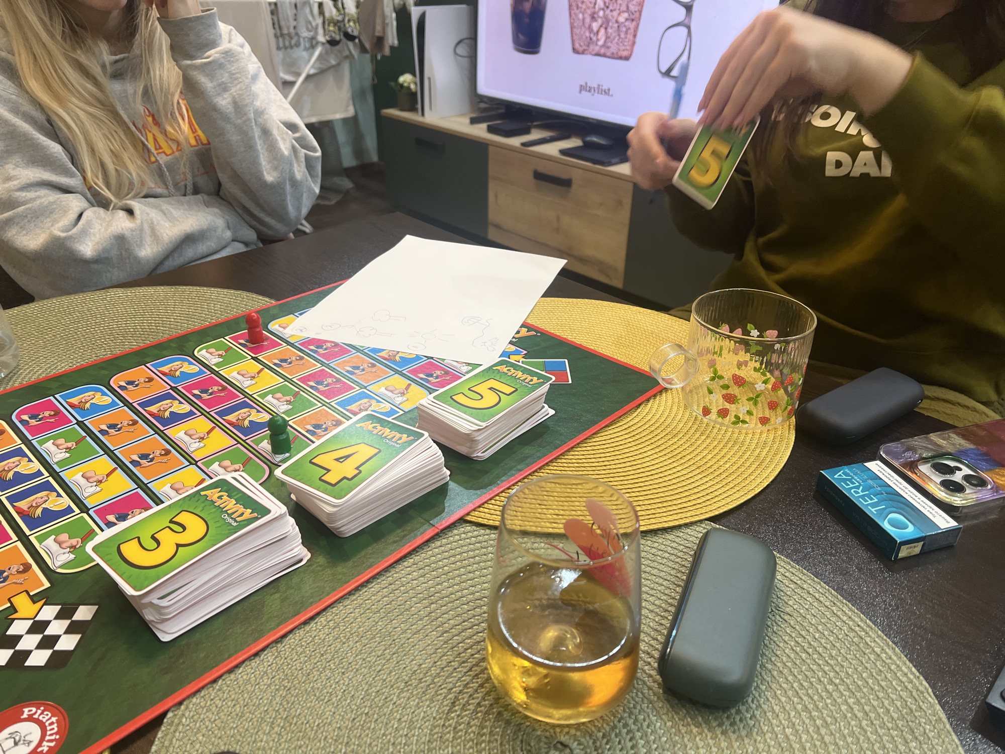 The image depicts a scene of people playing the "Activity" board game at a table. Two people are visible: one in a grey sweatshirt on the left and another in a green sweatshirt on the right, who is holding a "5" card. The game board is prominently displayed in the center of the image, along with piles of "Activity" cards numbered "3," "4," and "5." Two game pieces, one red and one green, are on the board. A white sheet of paper with sketches on it lies next to the board. In the background, there is a TV displaying a playlist and a PlayStation 5 console. On the table, there are two glasses with beverages, a smartphone, a package of TEREA tobacco sticks, and two grey rectangular devices. The location is not specified and cannot be inferred from the image.