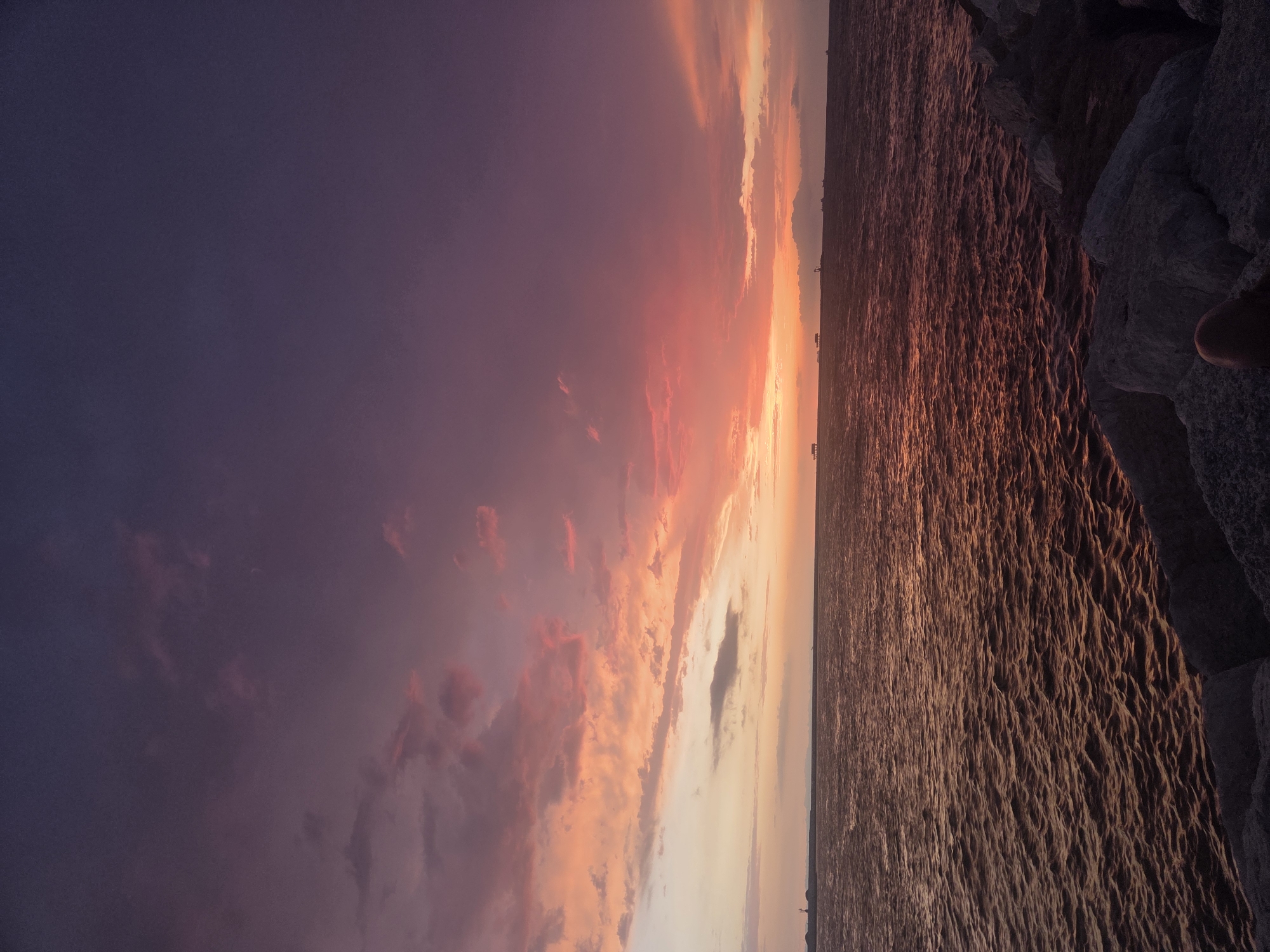 The image depicts a sunset over the Adriatic Sea, with the horizon visible and several structures and boats in the distance. The sky is filled with a dramatic display of color, transitioning from deep purple at the top to vibrant oranges and pinks closer to the horizon, indicating the sun has recently set or is about to set. Clouds are scattered across the sky, catching the light and contributing to the colorful spectacle. The sea reflects the colors of the sky, creating a shimmering, textured surface. In the foreground, large rocks are visible, suggesting a breakwater or shoreline. The scene is tranquil and evocative of a coastal sunset. While the image is of a coastal sunset, no specific landmarks or details definitively place it in Chioggia, Italy.