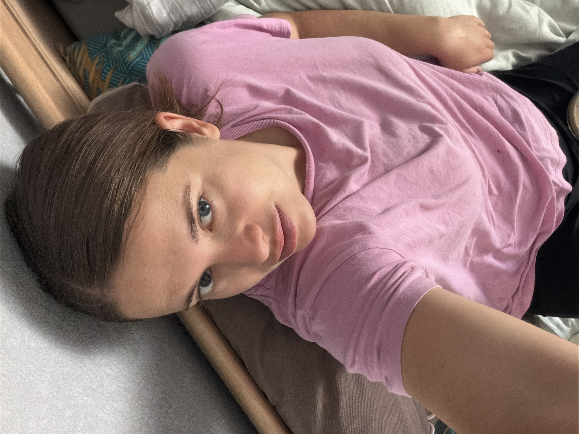 A young woman is captured in a diagonal, close-up selfie, lying down in an indoor setting. She has light brown hair, styled away from her face, and piercing blue eyes that look directly at the camera. Her complexion is fair, and her lips are slightly parted. She is wearing a casual, plain pink short-sleeved t-shirt and dark pants, a portion of which is visible on the right side of the frame. Her right arm, likely extended to hold the camera, is visible in the foreground.
The woman is resting on what appears to be a bed or couch, surrounded by pillows and bedding. To her immediate left, a light-colored, textured fabric, possibly grey or white, fills part of the frame. Behind her head, a pillow with a distinct teal, orange, and white geometric pattern provides a colorful accent. A plainer brown or grey cushion supports her head and upper body. A light brown, possibly wooden or upholstered, edge or headboard runs along the left side of the image.
The lighting is soft and diffused, suggesting natural daylight, likely morning or afternoon, filtering indoors. The absence of harsh shadows contributes to a relaxed and tranquil atmosphere. Her expression is calm and neutral, with a direct and engaging gaze. No visible text appears in the image. The overall environment is domestic and comfortable. The provided location context is Sochi, Russia.