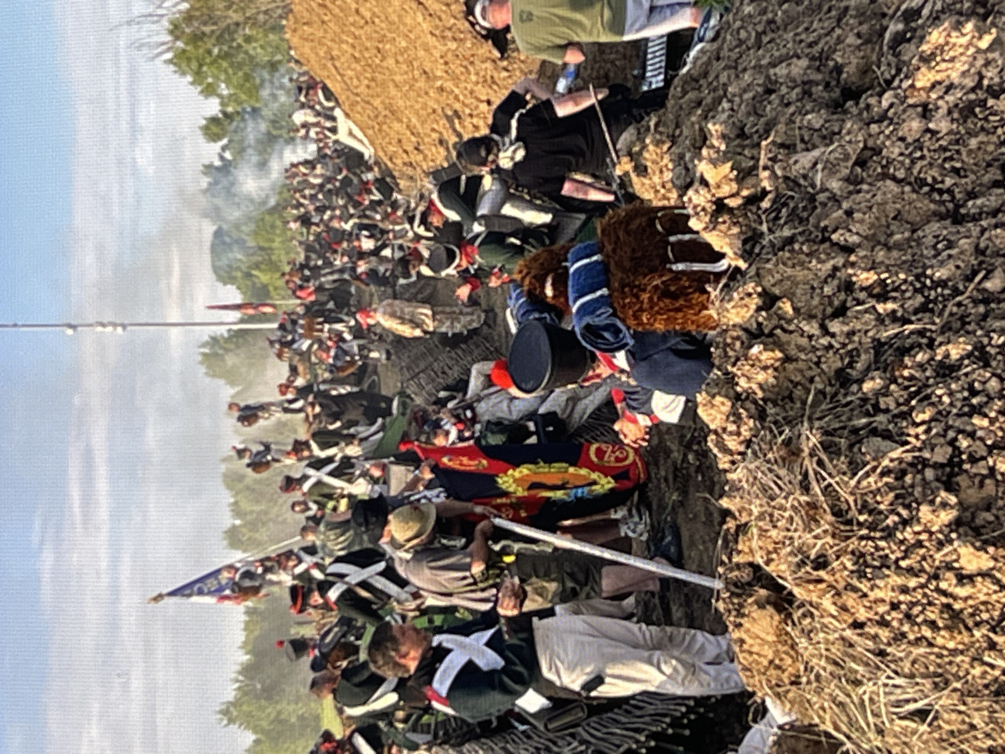 The image depicts a historical reenactment event. A large group of people dressed in 19th-century military uniforms are gathered outdoors on a sunny day. Many are wearing tall hats, some with plumes, and some are carrying flags and staffs. The foreground shows a pile of dirt and what appears to be a makeshift fortification. In the background, more participants are visible, some in formation, and there is a plume of smoke rising, suggesting a staged battle or cannon fire. The overall scene evokes a sense of a historical battlefield. The image is set in Moscow, Russia.