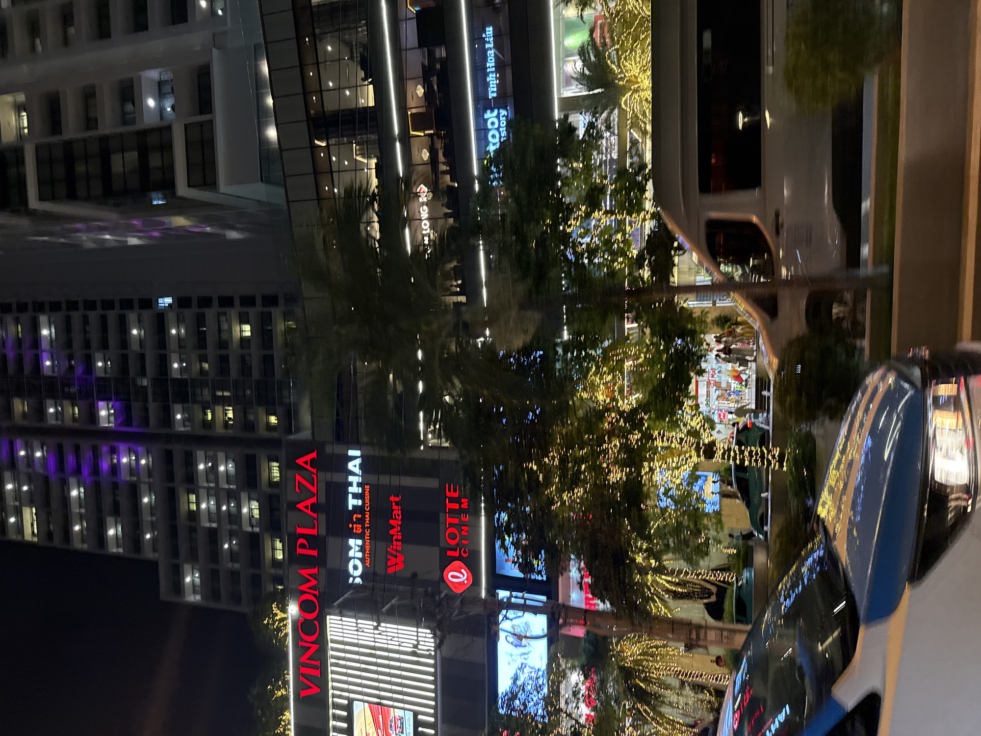 The image captures a night scene in Nha Trang Ward, Vietnam, focusing on the illuminated facade of Vincom Plaza. A large, modern building with numerous lit windows dominates the background. The Vincom Plaza sign is prominently displayed in red, along with other visible signs for "SOM THAI AUTHENTIC THAI CUISINE," "WinMart," and "LOTTE CINEM." Below these, illuminated screens display various images, and the building's exterior is adorned with strings of bright, twinkling lights, particularly around trees and balconies, creating a festive atmosphere. In the foreground, the front of a white car, partially visible with its headlights on, suggests movement. A white van is also seen in motion to the right, passing by the plaza. Several individuals are present, some walking in front of the plaza's entrance and along pathways, while others are inside visible through the windows of the building. The overall environment is urban and commercial, with a focus on the bright, vibrant displays of the shopping and entertainment complex at night.