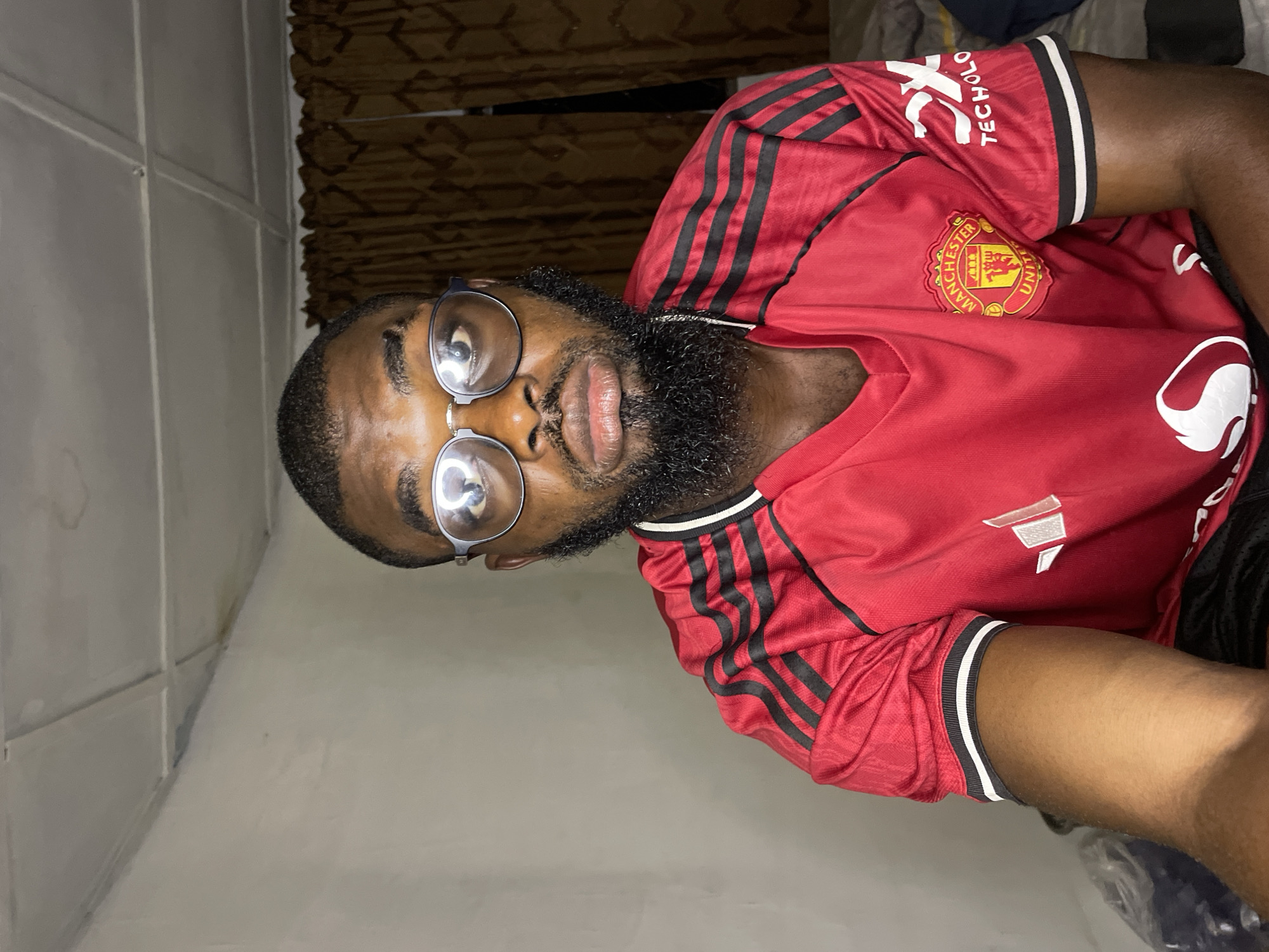 A close-up, head-and-shoulders portrait of a Black man wearing glasses and a red Manchester United jersey is presented. The man has a beard and appears to be looking directly at the camera with a neutral expression. The background consists of a plain, light-colored wall and brown curtains. The lighting suggests an indoor setting, possibly a bedroom. The image does not contain any specific elements that would allow for the identification of its location within Jos, Nigeria.