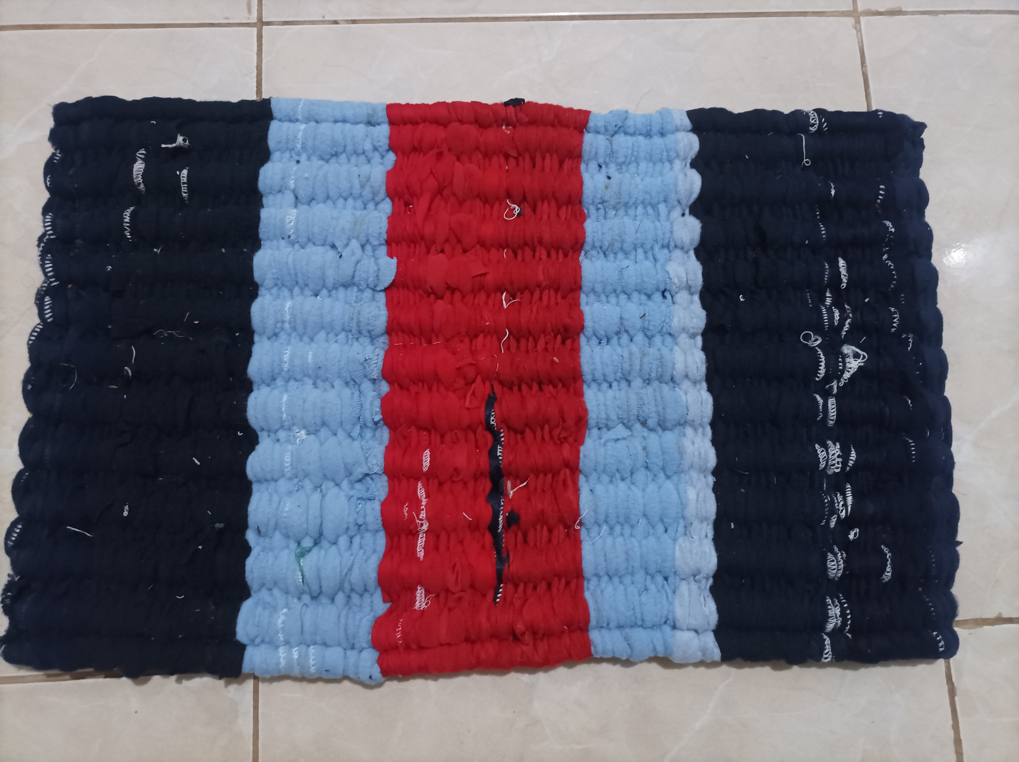 The image displays a rectangular woven rug lying on a tiled floor. The rug is divided into three vertical sections of color: black on the left, light blue in the middle, and red in the center. The black and light blue sections are repeated, creating a pattern of black, light blue, red, light blue, and black. The rug appears to be handmade, with a slightly uneven texture. The tiles beneath the rug are beige and arranged in a grid pattern. The image is taken from a top-down perspective. There is no visible indication of location within Nganjuk, Indonesia.
