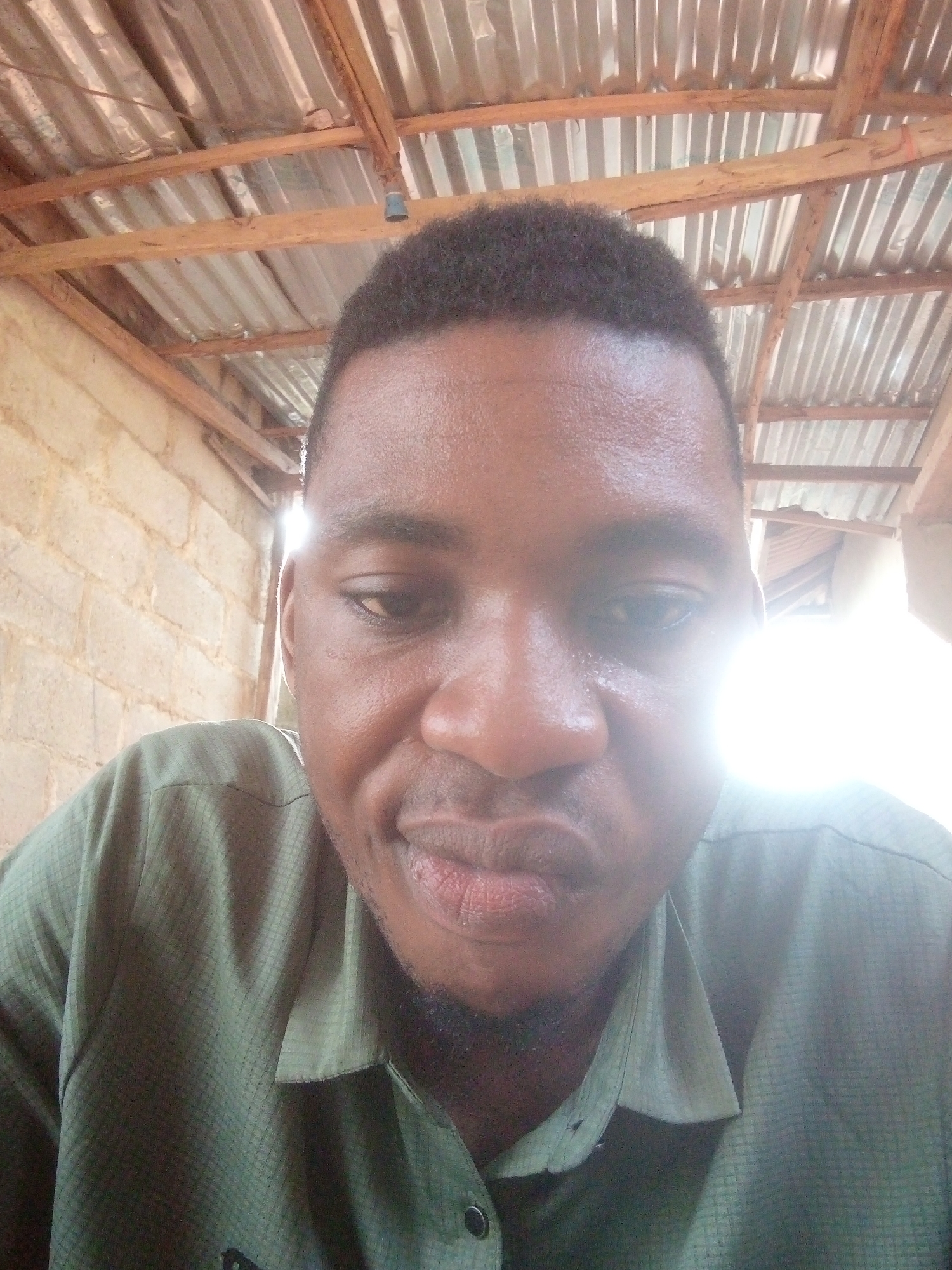 A close-up selfie of a Black man with a short haircut and a slight beard is set against a background indicating a construction or outdoor shelter. He is wearing a dark green shirt. The background reveals a roughly constructed ceiling made of wooden beams and corrugated metal. To the left, a wall made of concrete blocks is visible. The location is in the city of Unknown, Unknown. A bright light source, possibly the sun, is visible in the background on the right side of the frame.