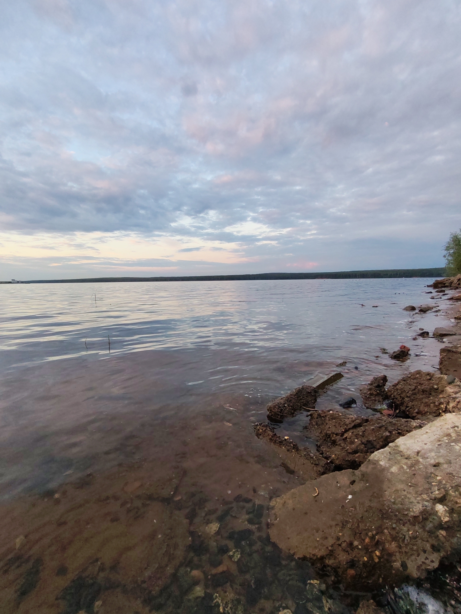 The image depicts a serene natural scene of a body of water, likely a river or lake, with a treelined horizon under a cloudy sky. The foreground features a rocky shoreline with scattered debris, including concrete fragments and what appear to be natural rocks. The water is relatively calm, reflecting the soft colors of the sky, which shows hues of blue, grey, and light pink. The depth of the water near the shore is shallow, revealing the rocky bottom. The overall atmosphere is tranquil and suggests an undeveloped or natural area. The location is identified as Chaikovsky, Russia, which is known for its natural beauty and proximity to the Kama River.