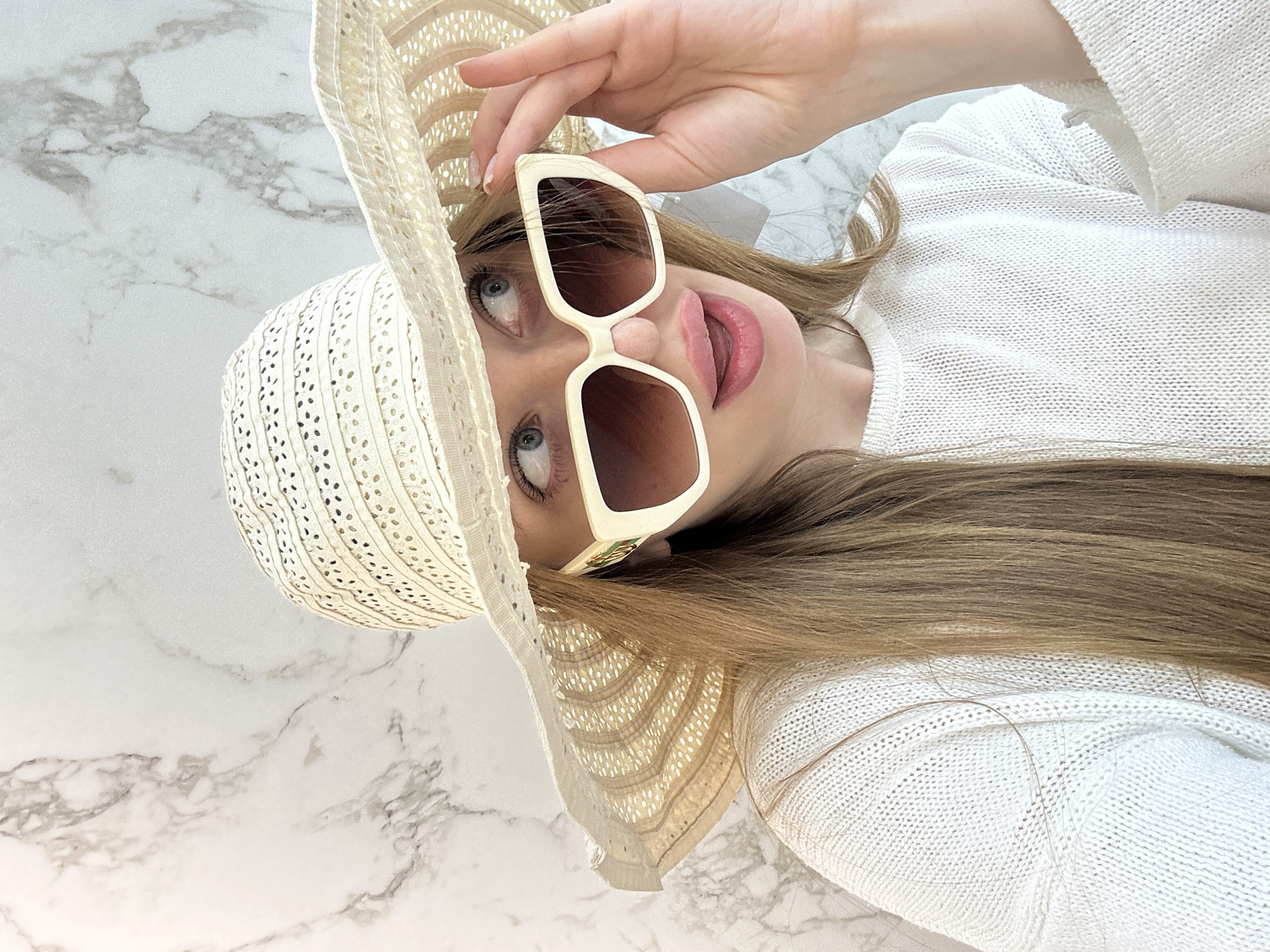 A light-skinned woman wearing a white sweater, a beige straw sunhat, and off-white sunglasses is visible in a portrait-style photograph taken indoors. The background appears to be a marble surface. The photograph is vertically oriented and taken from an unusual, tilted angle, so the woman is rotated counter-clockwise by roughly 90 degrees. No specific details allow for the identification of Chaikovsky, Russia.