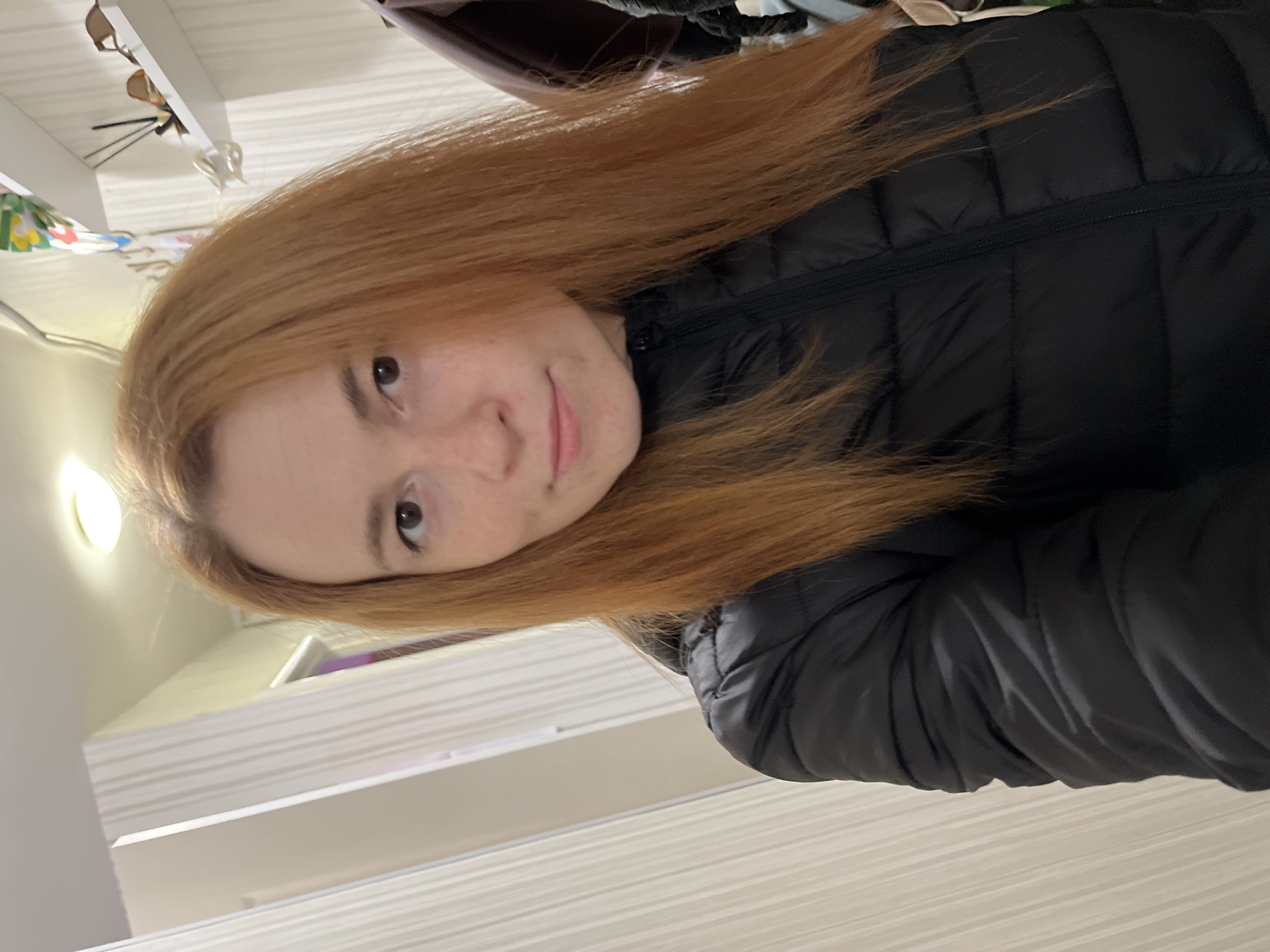 A woman with light skin and red hair wearing a black puffer jacket is pictured indoors. The location is unknown.