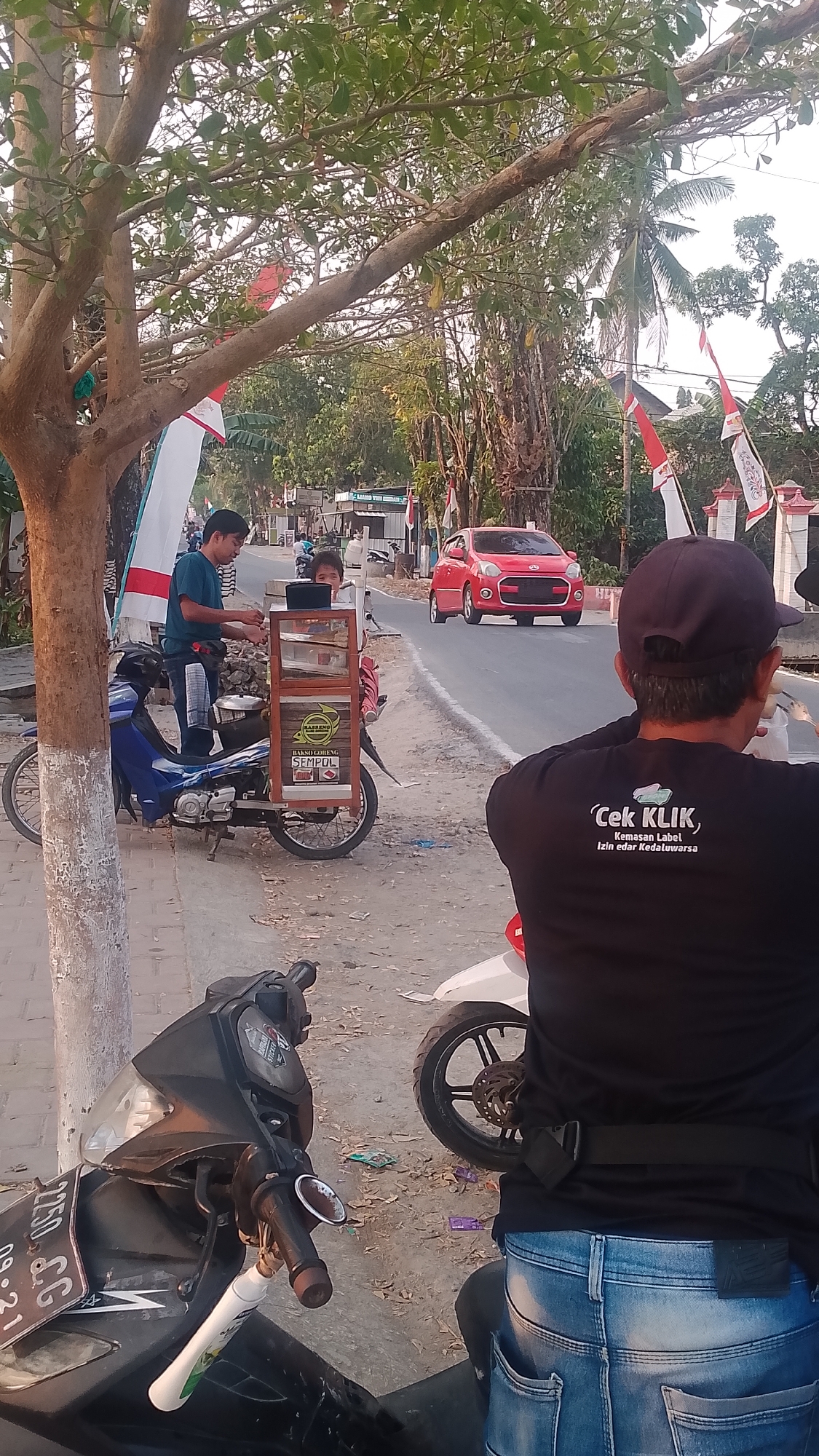 In Karanglangu, Indonesia, a roadside scene unfolds under the shade of a leafy tree. Two men and a child are near a food cart mounted on a blue motorcycle, which is parked on a dirt shoulder. One man, wearing a dark blue t-shirt and jeans, stands by the cart, while the other, in a black t-shirt and jeans, sits on a white motorcycle in the foreground, facing away from the camera. A child with dark hair observes the cart. The food cart displays "Bakso Goreng Sempul" and has a glass display case. Behind them, a red car drives on a paved road lined with trees and Indonesian flags. The environment appears to be a suburban or rural road with sparse vegetation and a few buildings in the distance.