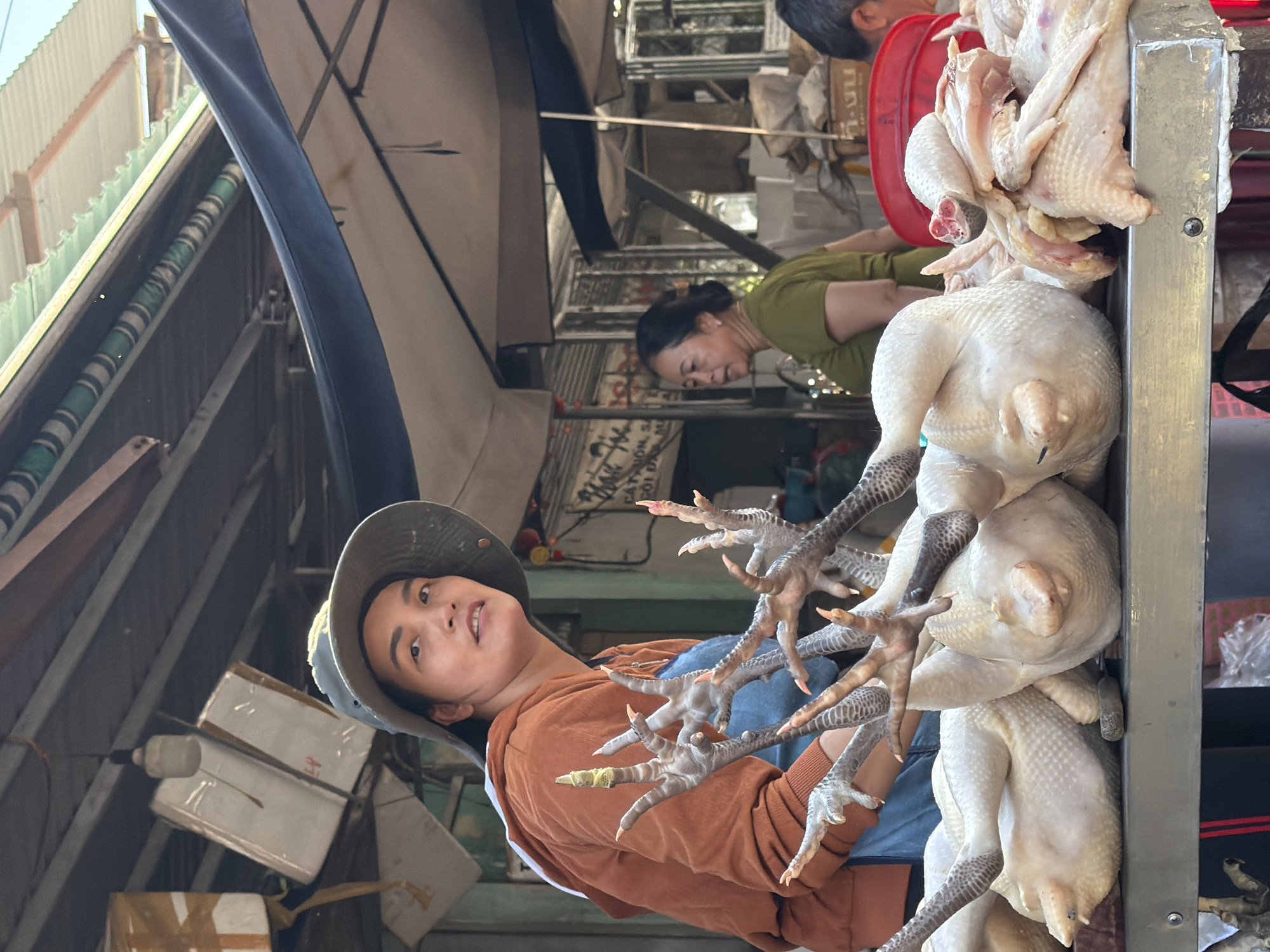 A woman wearing a wide-brimmed hat and an orange hooded sweatshirt stands behind a display of whole chickens and chicken parts at an outdoor market in Nha Trang, Vietnam. She is looking towards the camera with a neutral expression. Several raw chickens are laid out on a metal table in front of her, their legs prominently displayed. Behind her, another woman in a green shirt is partially visible, and a red plastic tub is present. The market appears to be covered by awnings and corrugated metal roofing. The overall scene depicts a typical street market environment where fresh produce, including poultry, is sold.