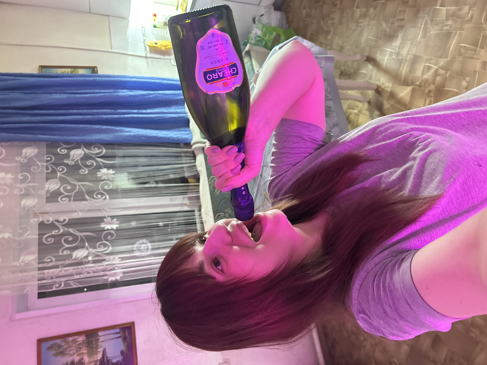 A woman holds a bottle of CHEARO Blanco towards her mouth. She is indoors, in front of a window with patterned blinds and blue curtains. Two framed pictures are visible on the wall. The lighting appears to have a pink hue. The bottle is green with a label. The location cannot be inferred as Фоки, Russia without more context.