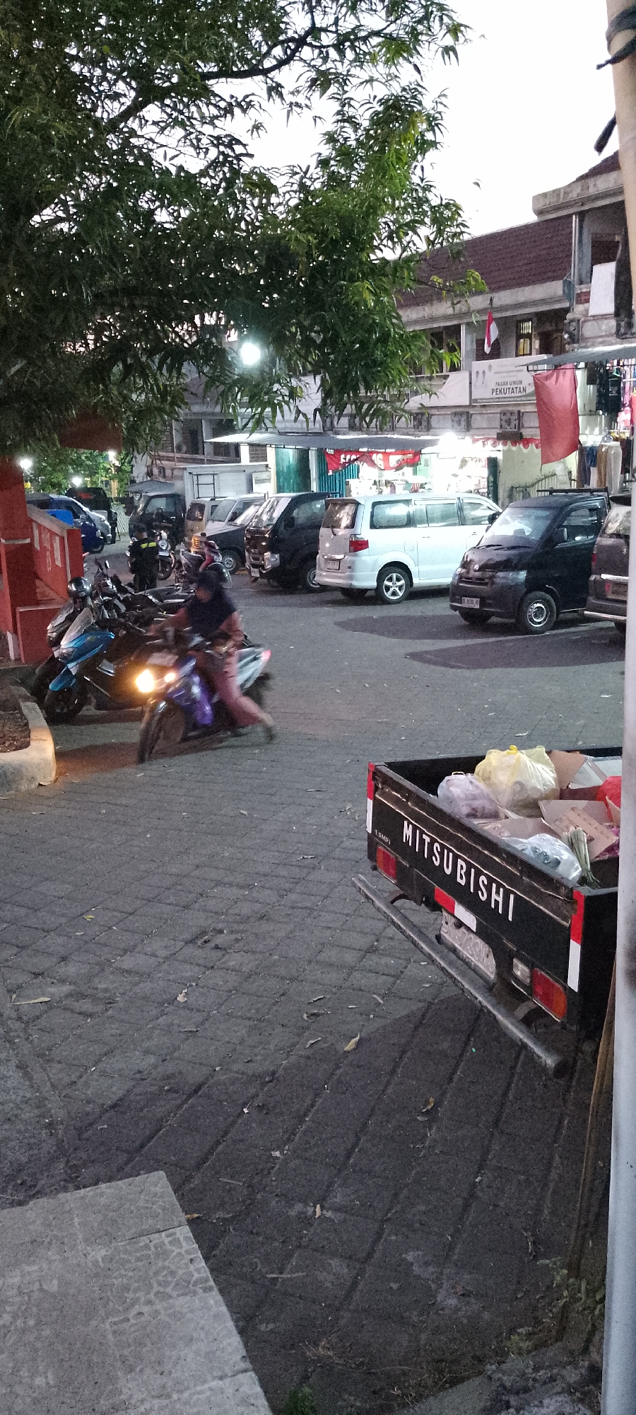 The image depicts an outdoor scene in Pekutatan, Indonesia, likely a market or parking area, during twilight. A dark-colored Mitsubishi pickup truck is parked on the right side of the frame, its bed filled with various items including plastic bags and cardboard boxes. In the foreground, a person is riding a light-colored motorcycle with its headlight illuminated, moving from left to right across the paved ground. To the left, several other motorcycles are parked. In the background, a building with a red roof and a sign that reads "PASAR UMUM PEKUTATAN" (Pekutatan Public Market) is visible. Trees with green leaves overhang parts of the scene. The ground is paved with grey interlocking bricks. The overall lighting suggests a low-light condition, possibly early morning or late evening.