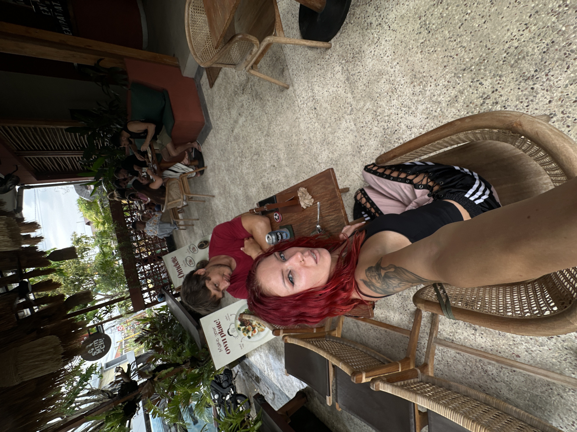 A woman with bright red hair is taking a selfie in the foreground, her arm extended towards the camera. She is wearing a black tank top and pink pants with black lace-up detailing and white stripes. Behind her, a man in a red t-shirt is seated at a wooden table, looking down. A menu board is propped up on the table, displaying "Make your own plate!" and "Omelette" with illustrations of food. In the background, other patrons are seated at tables, some eating and others conversing. The setting appears to be an outdoor cafe or restaurant with a thatched roof overhead, lush greenery, and rattan chairs and tables. The floor is a speckled concrete. A sign that reads "Munch-Chi" is visible hanging from the thatched roof structure. The overall scene suggests a casual dining experience in North Kuta, Indonesia.