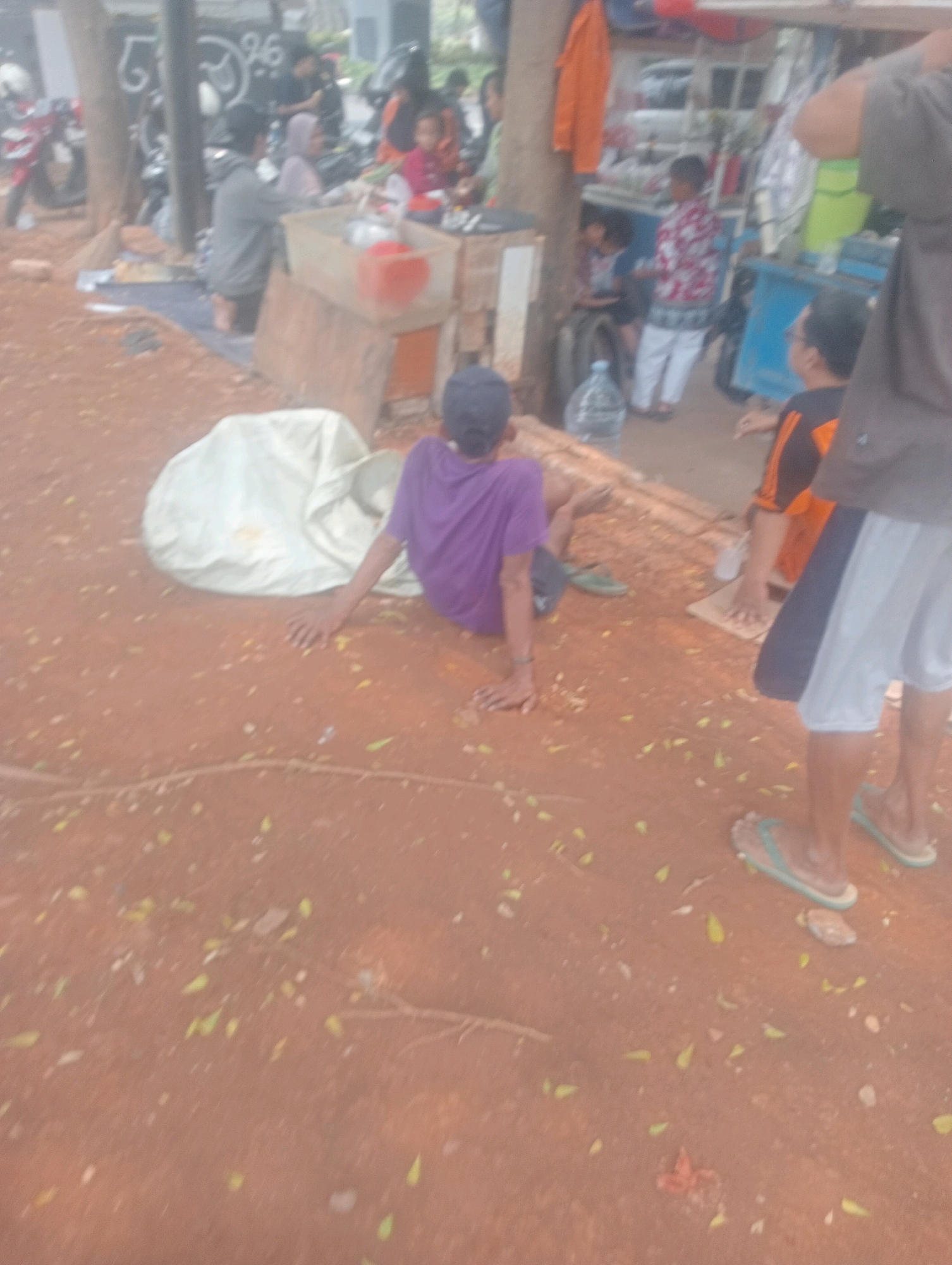 The image shows an outdoor scene with several people in what appears to be an informal settlement or market area in South Jakarta, Indonesia. In the foreground, a young person wearing a purple t-shirt is sitting on the ground with their legs extended. Next to them, to the right, another person in an orange and black shirt is also seated, with their hands near a piece of cardboard on the ground. In the background, several other individuals are visible, some standing and some sitting, engaged in various activities.
To the left of the seated figures, a large white object, possibly a tarp or a bag, is visible. The ground is covered in reddish-brown soil with scattered green leaves. In the background, there are structures that suggest temporary stalls or shelters, with some people gathered around them. There are also motorcycles parked in the vicinity, hinting at a busy, communal environment. The overall impression is a snapshot of daily life in a densely populated urban area in South Jakarta, characterized by informal commerce and social interaction.
