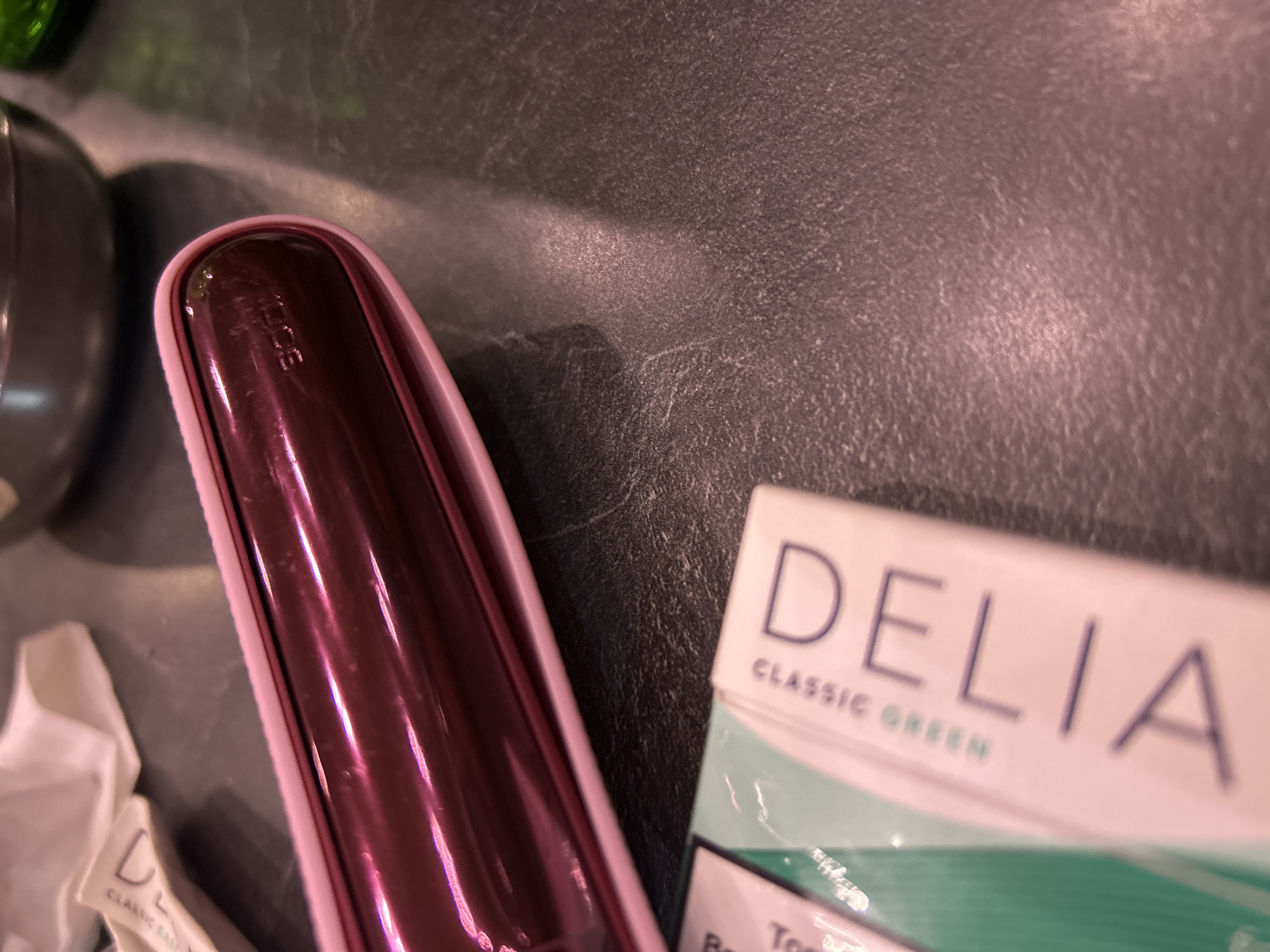 The image presents a close-up, angled view of personal items arranged on a dark, textured surface, likely indoors in Plovdiv, Bulgaria.
The primary subject is a sleek, elongated electronic device positioned on the left side of the frame. It boasts a glossy, metallic pinkish-purple finish with a rounded top end. Faint, embossed lettering "iQOS" is visible near this end, identifying it as an iQOS tobacco heating device.
Adjacent to the iQOS device, on the right, a white and green product box is partially visible. The box features prominent dark lettering spelling "DELIA" above the phrase "CLASSIC GREEN." Below this, a faint "Too" is discernible, suggesting the product might be related to toothpaste or another personal care item. In the bottom-left corner, a crumpled piece of white fabric or paper, possibly a wipe, shows partial text including the letter "D" and "CLASSIC," indicating it may be associated with the "DELIA CLASSIC" brand.
The background consists of a dark, subtly textured surface, resembling a countertop or table. A dark, rounded object, possibly a container or part of another item, is partially visible in the top-left corner. The lighting appears to be indoor, and no specific activity or emotions are depicted. The scene is a static arrangement of objects.