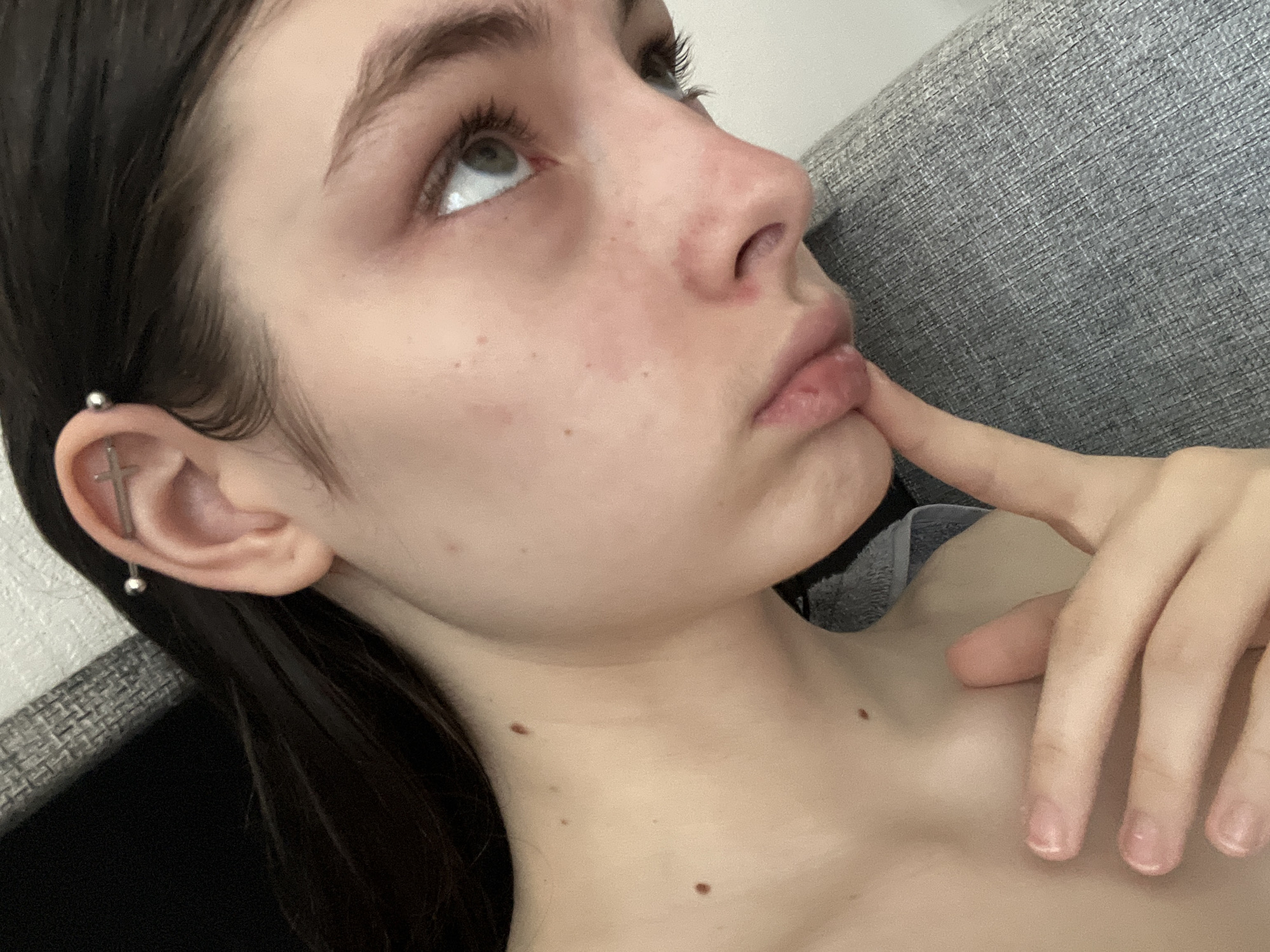 A young, light-skinned individual with dark hair and visible moles is positioned against a gray, textured background, possibly a sofa or other upholstered surface. The person's head is tilted upwards and to the left, with their eyes looking in the same direction. A finger is positioned near their lips, suggesting a contemplative or pensive mood. The individual has a distinctive ear piercing resembling a cross. The background is out of focus, but appears to be a neutrally decorated indoor space. There is no evidence to determine whether this is Saint Petersburg or any other location.