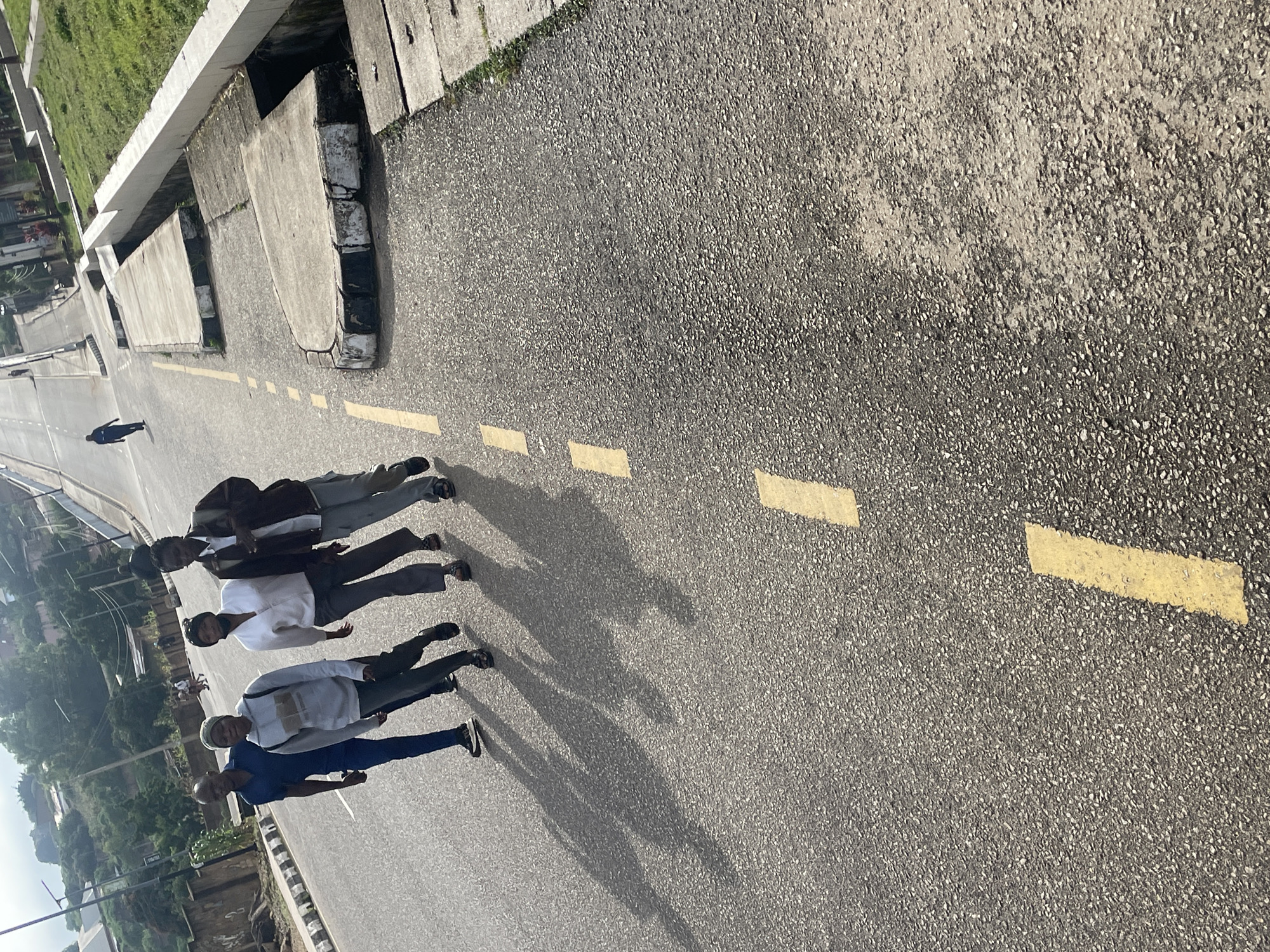 In Jos, Nigeria, four individuals are walking on an asphalt road. The group consists of three women and one man. The man, on the far left, is wearing a blue polo shirt and dark trousers. Next to him is a woman in a grey hooded sweatshirt and dark trousers. To her right is another woman dressed in a white button-down shirt and grey trousers. The last person in the group, on the far right, is a woman wearing a dark jacket over a lighter-colored shirt and grey trousers. The road has painted yellow lines, indicating traffic lanes. In the background, there are buildings, trees, and a hillside. The sun casts long shadows of the individuals onto the road, indicating it is likely morning or late afternoon.