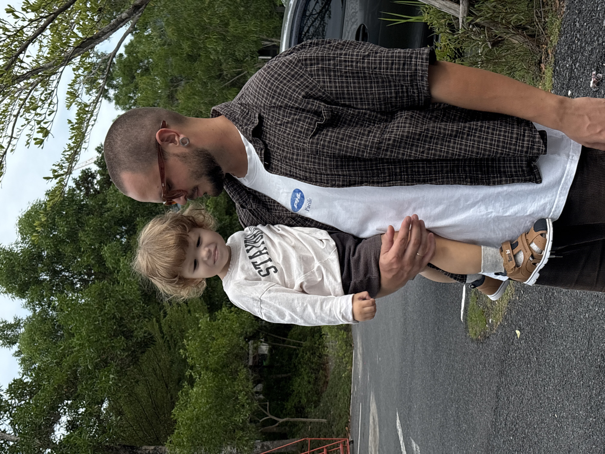 A man with short hair, a beard, and sunglasses holds a young child in his arms. The man wears a white t-shirt, a brown plaid button-down shirt, and dark pants. The child wears a white long-sleeved shirt with black lettering, dark shorts, and brown sandals with white socks. They are standing on an asphalt road next to greenery, trees, and a black vehicle. The image appears to be taken outdoors, potentially in Choeng Thale, Thailand, based on the query context.