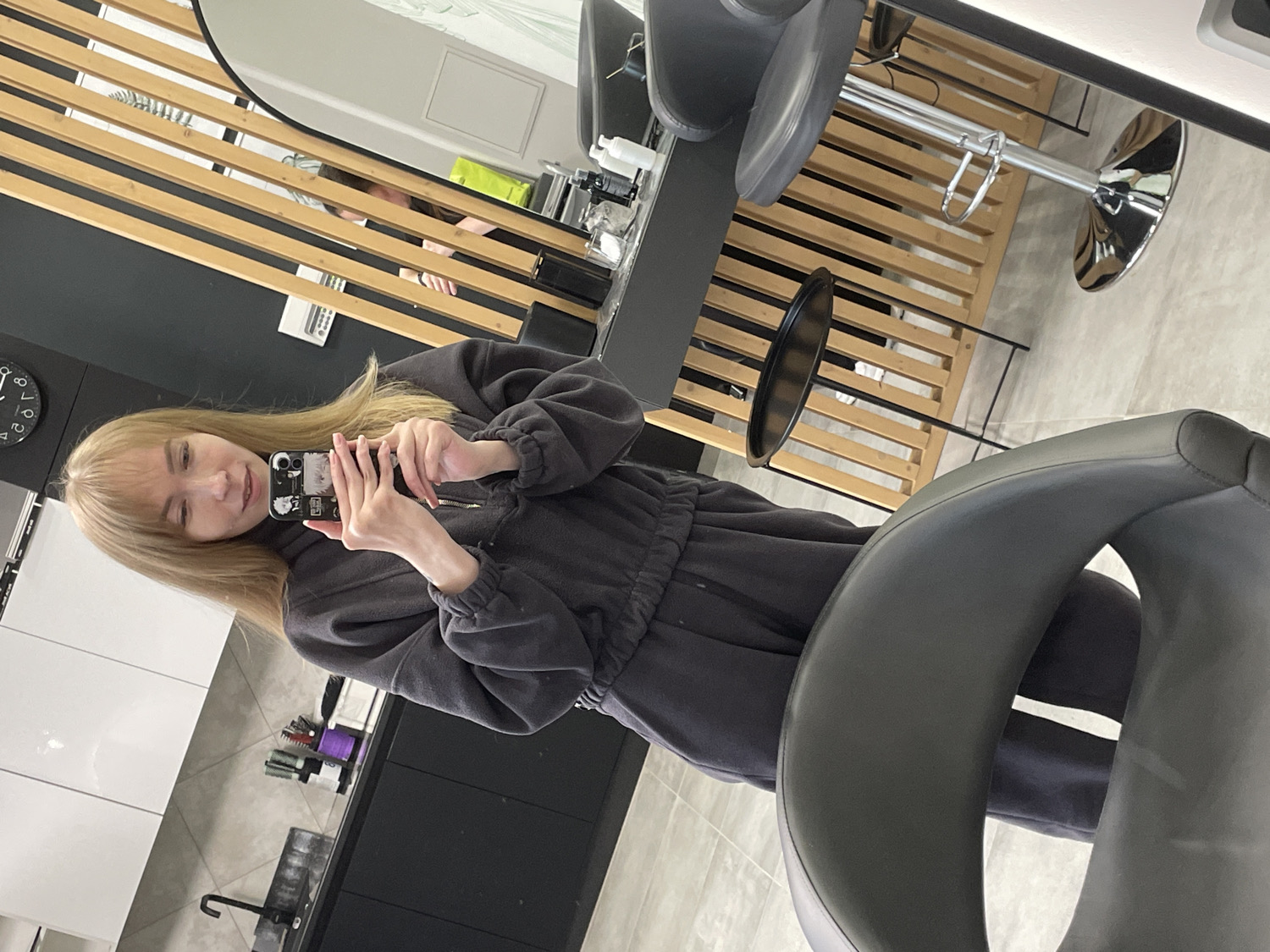 The image, taken with a tilted perspective, shows a young woman capturing a selfie in what appears to be a modern hair salon or beauty studio. She has long, straight blonde hair with bangs and a fair complexion, and is smiling faintly. She is wearing a dark grey, possibly charcoal-colored, casual tracksuit, comprising a zippered hoodie and matching sweatpants. Her smartphone, used for the selfie, has a black case adorned with a few white stickers or graphics.
The background reveals a sleek and contemporary interior. A large, oval-shaped mirror behind the woman reflects a wooden slatted room divider, and partially, another person with dark hair, possibly a stylist or another client. To the left of the frame, a black wall features a circular clock with white numbers (1 through 12 visible, though rotated due to the image tilt), with its hands indicating a time approximately between 2:45 and 3:45 if oriented upright. Below the clock, there are white upper cabinets above a dark countertop fitted with a sink and a black faucet. Various salon tools, such as brushes and product bottles, are neatly arranged on the counter.
In the immediate foreground, part of a dark grey, upholstered chair, likely a salon chair or a bar stool, is visible. The floor is covered with light grey tiles. The overall scene suggests a clean, well-lit, and professional environment. Based on the provided context, the location is Perm, Russia. The woman's expression conveys a sense of calm contentment.