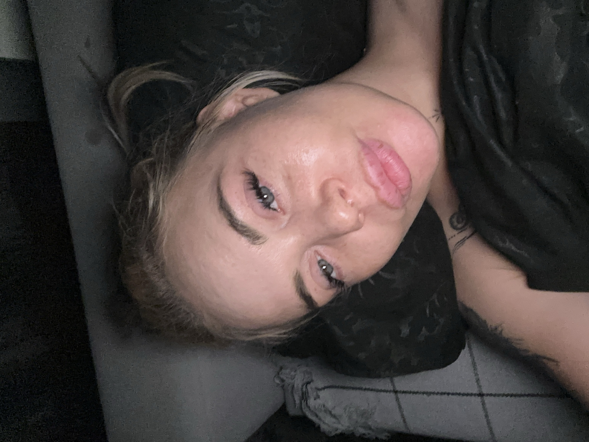 The image captures a close-up, high-angle portrait of a female subject lying down in an indoor setting in the city of Unknown, Unknown. The subject's head and upper torso are visible, with her gaze directed towards the camera. Her hair, light brown with blonde highlights, is pulled back into an untidy bun or ponytail, with loose strands around her face. Her eyes are partially open, featuring long, dark eyelashes. Her lips are full, and her skin exhibits a glossy texture on the forehead and cheeks. A small, dark tattoo is visible on the inner right forearm, and a faint, small tattoo is present on the lower neck/upper chest area. The subject is partially covered by a dark, soft-textured blanket or duvet. To the left, a section of a lighter-colored, possibly checkered or plaid, fabric object is visible, likely a blanket or pillow. The background is dark and indistinct.