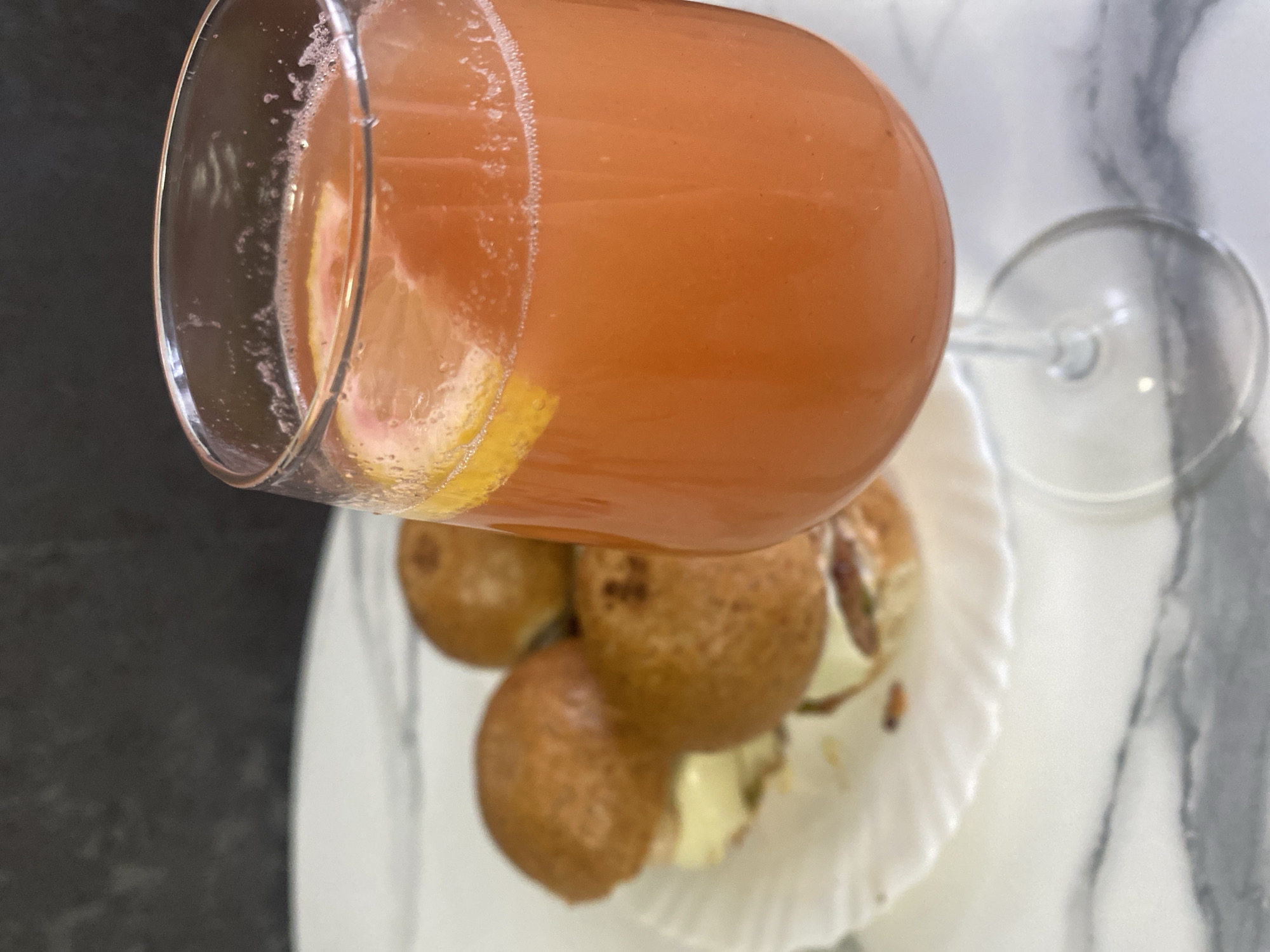 The image shows a tilted champagne flute filled with a pink cocktail, containing a slice of citrus, likely grapefruit or lemon. Below the glass is a white, ruffled-edge plate with a cluster of small, round pastries, possibly filled with cream. The background surface is a white marble pattern on the right and a dark, solid surface on the left. The setting and details in the image are not sufficient to determine if the location is in the city of Izhevsk, Russia.