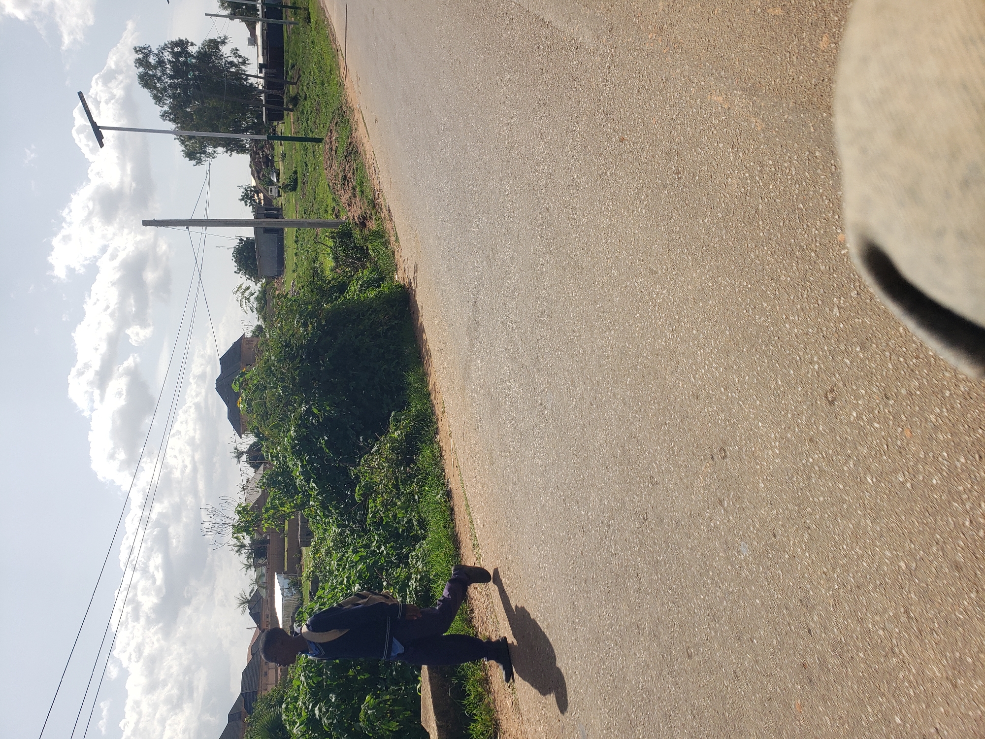 A person walks on the right side of a paved road in Gura Top, Nigeria. The person is wearing dark pants, a dark long-sleeved shirt, and black shoes. A backpack with light-colored straps is worn on their back. The person's left leg is extended forward as they walk. Sunlight casts a shadow of the person on the road to their left. Lush green foliage lines the left side of the road, featuring dense bushes and some taller trees. In the background, several residential buildings with dark-tiled roofs are visible, interspersed with smaller structures. Power lines crisscross the sky. A tall lamp post with solar panels stands on the right side of the road. The sky is partly cloudy with white cumulus clouds.