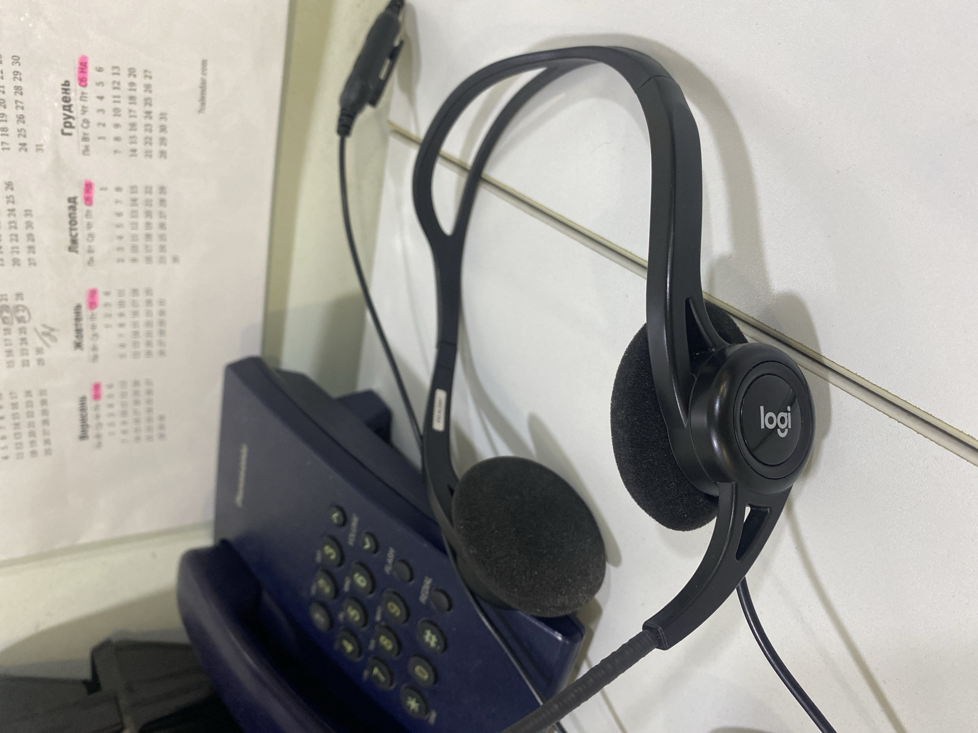The image depicts a close-up, overhead view of a Logitech headset resting on a white, tiled surface. The headset is black with two earcups and a flexible headband. To the left of the headset is a dark blue corded telephone with a handset lying on its side. Behind the telephone, a portion of a calendar is visible, showing dates and days of the week in Cyrillic script. The calendar indicates the months of November and December. The overall scene suggests an office or home office environment. The image does not provide any information about the city of Rozhny, Ukraine, or any specific activities taking place.