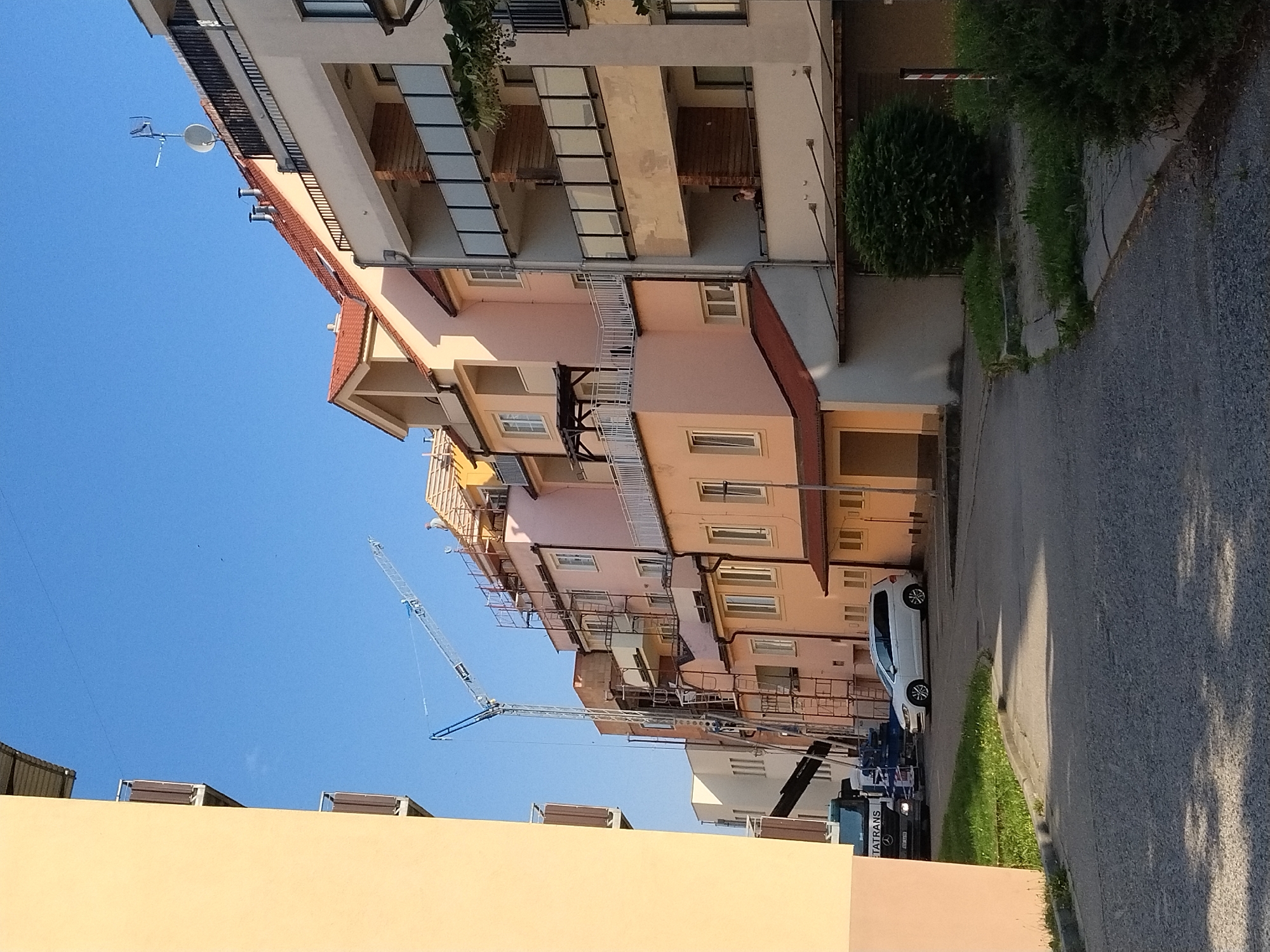 In Hluboká nad Vltavou, Czechia, on a sunny day, a street scene unfolds with residential buildings and construction activity. A tall construction crane is prominently positioned, its jib extending over a multi-story apartment complex that is undergoing renovations or expansion, indicated by scaffolding and exposed building materials. A white minivan is parked on the asphalted road in front of the buildings. Adjacent to the road, a strip of grass runs along a curb. To the left, a portion of a yellow-hued building with balconies is visible, partially obscuring the view of the construction site. The sky is clear and blue.