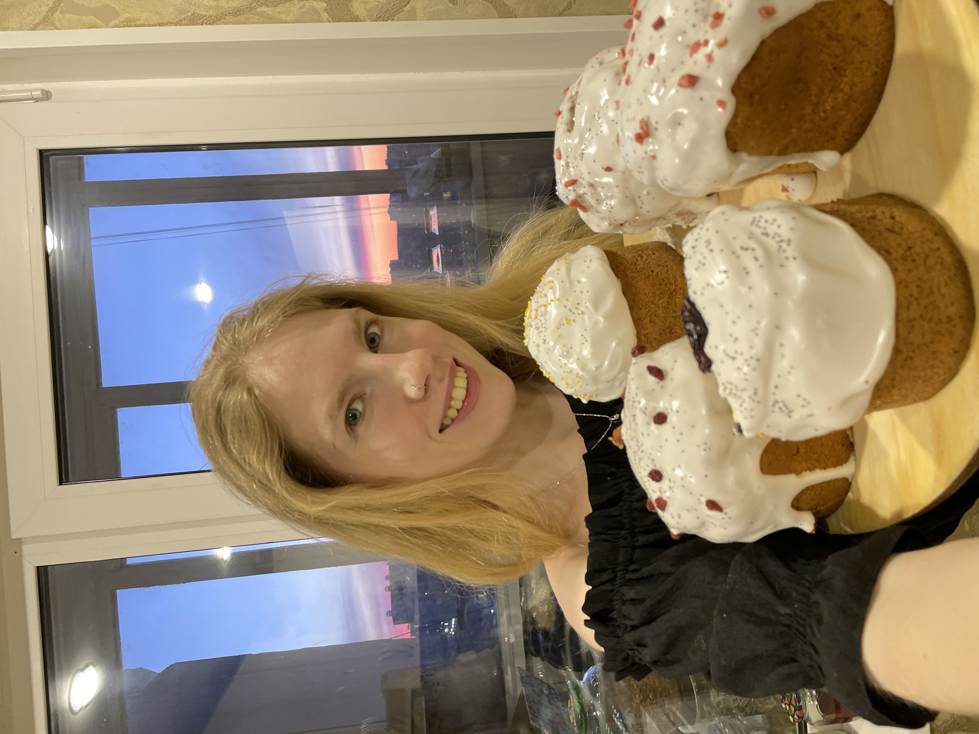 A person with blond hair, wearing a black top, is holding a plate of decorated cakes. The cakes have white icing and various colored sprinkles. The scene is indoors, possibly in an apartment, with a window showing the Izhevsk cityscape at dusk. The window frame is white. The wallpaper behind the window frame is a light brown patterned design. The person is smiling and looking at the camera.