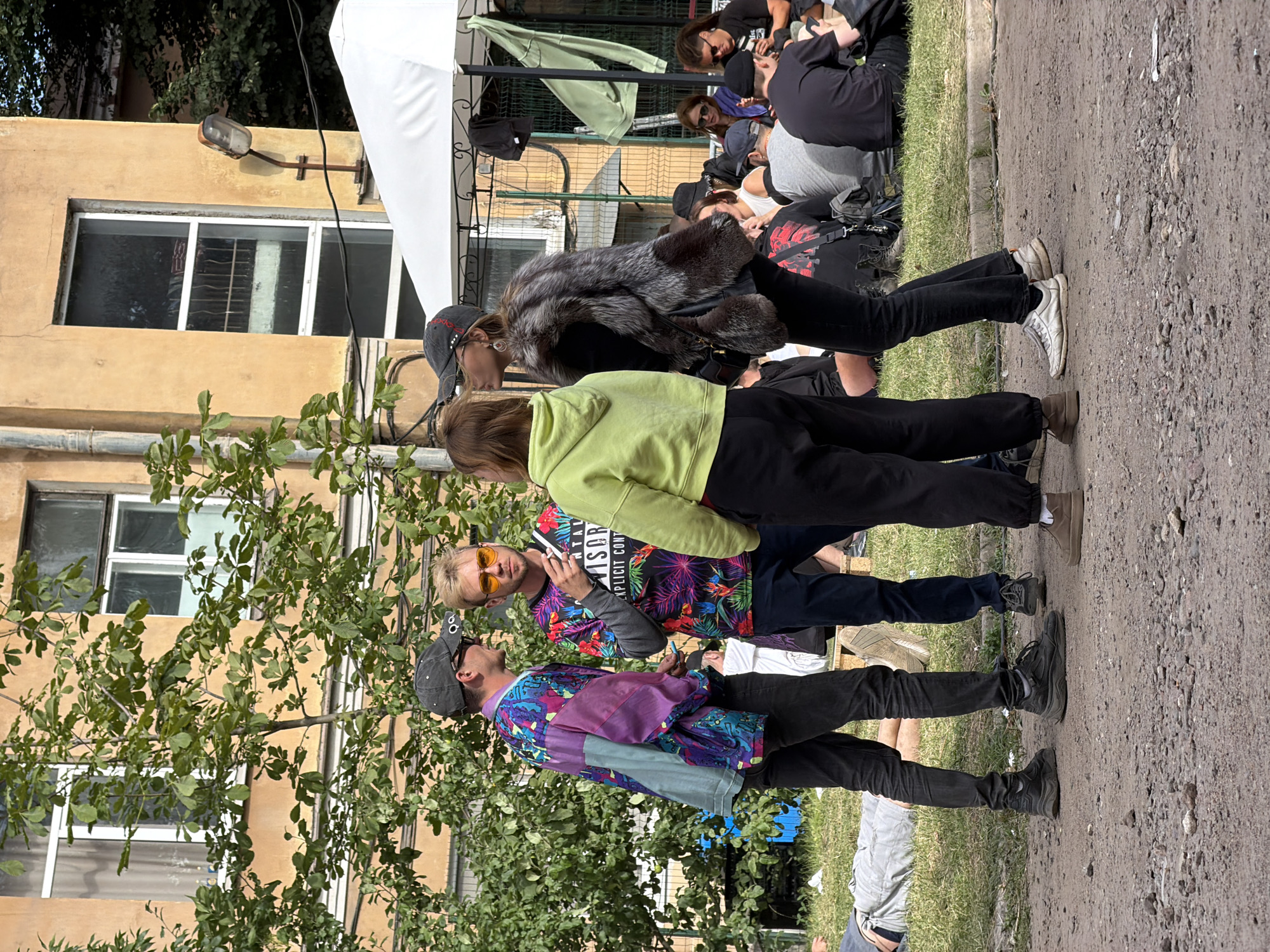 The media file displays a candid, outdoor scene of several people gathered on a sunny day, likely in Saint Petersburg, Russia. The image is oriented vertically. In the foreground, three young men are interacting. The man on the left, wearing a dark baseball cap and a vibrantly patterned jacket in shades of purple, blue, and green, appears to be holding something like a cigarette or a small object. He is looking at the man in the center. This central figure, sporting sunglasses and a tropical-patterned shirt with the visible text "EXPLICIT CONT", is engaged in a conversation, holding a phone to his ear. To the right of these two, a third person, wearing a light green hooded sweatshirt, is looking down.
Behind them, another group of people are seated on the ground, some appearing to be resting or socializing. One individual in the background wears a faux fur jacket. The setting includes a building with a yellow facade and windows, with a tree and green foliage in the left foreground. A white tent or awning structure is visible in the upper part of the image, suggesting a possible event or gathering. The ground is a mix of dirt, gravel, and grass. The overall atmosphere seems relaxed and casual. The time of day appears to be daytime due to the bright lighting.