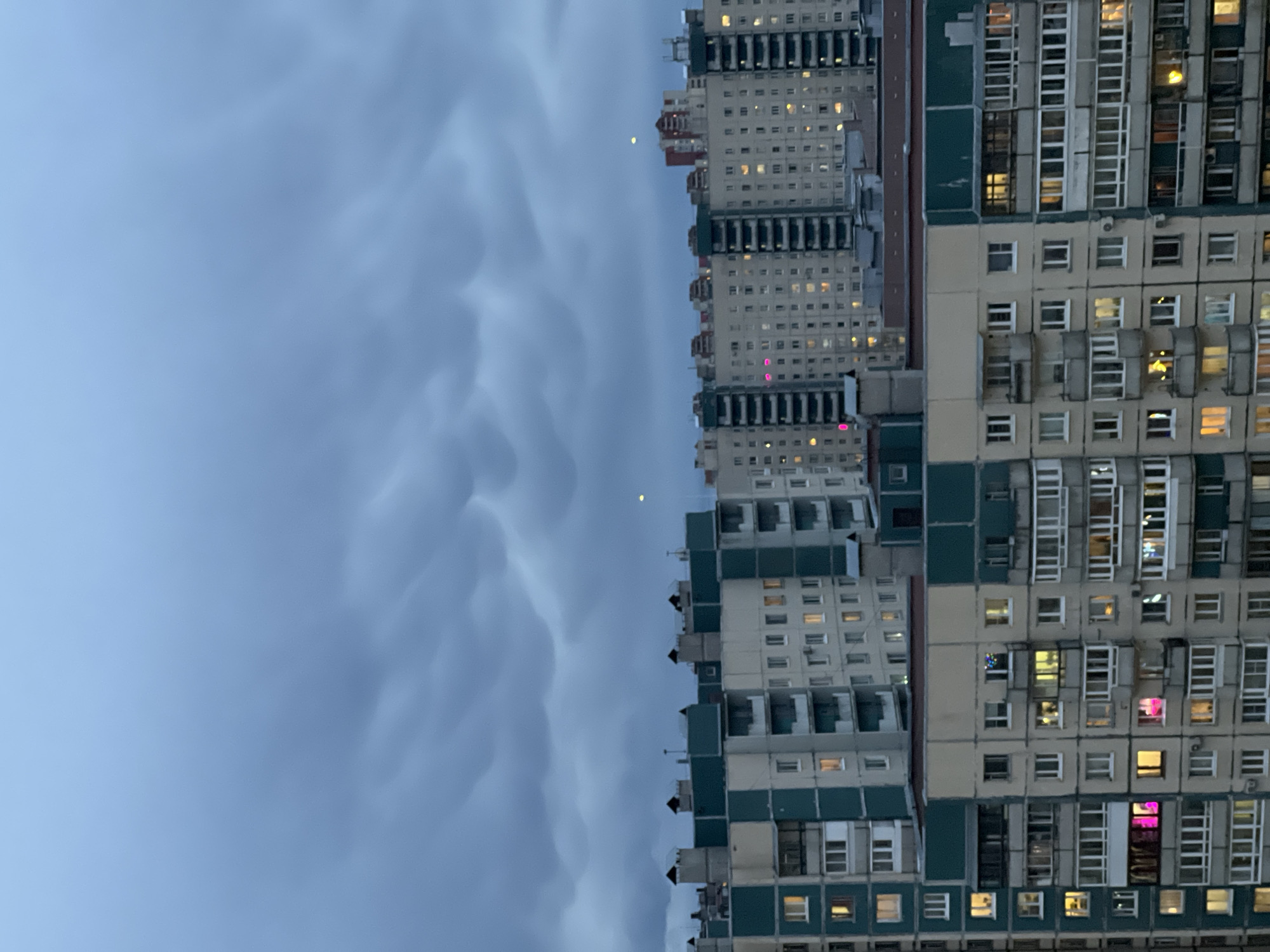 The image displays a towering urban residential scene at what appears to be twilight or night. On the right side, multiple large, multi-story apartment buildings dominate the frame, exhibiting a distinct architectural style consistent with post-Soviet construction, aligning with the provided Saint Petersburg context. These buildings feature facades in shades of light beige/grey and dark teal/green, with some reddish-brown accents visible on one structure. Numerous windows punctuate the buildings' surfaces, many of which are illuminated from within, suggesting active occupancy. The internal lights cast a warm glow, contrasting with the exterior darkness.
The left side of the image is filled with a dramatic sky. It is a deep blue-grey, indicative of a time between dusk and dawn. The sky is characterized by distinct, wavy cloud formations, possibly mammatus clouds, adding texture and depth to the celestial expanse. Two small, bright points of light are visible in the sky, potentially stars or distant artificial lights, given the low light conditions. No specific human activity is directly observable, but the lit windows imply residential life within the buildings. There is no visible text in the image.