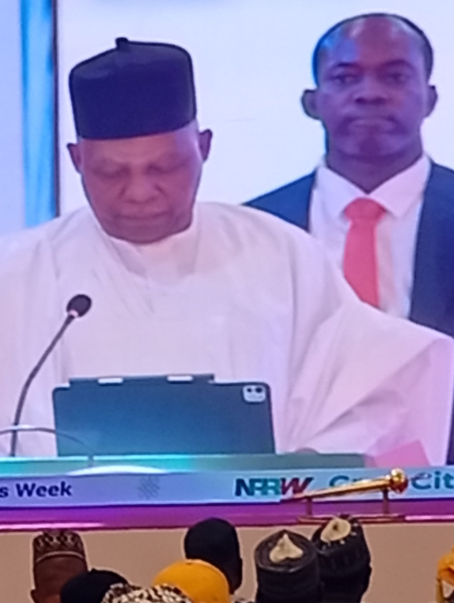 An indoor event is captured, featuring two prominent figures on a stage or platform. In the foreground, a man of African descent wears a white traditional attire, possibly a caftan or agbada, and a black square cap. He stands behind a podium with a microphone in front of him and a tablet device resting on the surface. His head is bowed, and his eyes appear closed or looking down, conveying a moment of focus or solemnity.
Standing slightly behind and to the right of the primary speaker is another man, also of African descent, dressed in a dark suit, a white shirt, and a bright red tie. He looks straight ahead with a neutral expression.
The lower part of the image shows the tops of the heads of an audience, many wearing traditional headwear, indicating a crowd observing the event. A banner runs across the front of the podium. Partially visible text on the banner includes "s Week" on the left and "NRBW C it" on the right. A golden, ornate ceremonial staff or scepter-like object rests horizontally on the banner on the right side. The setting is brightly lit, suggesting a formal gathering or conference during the daytime. The attire and general context align with events that might take place in Nigeria, as suggested by the Tudun Nupawa location.