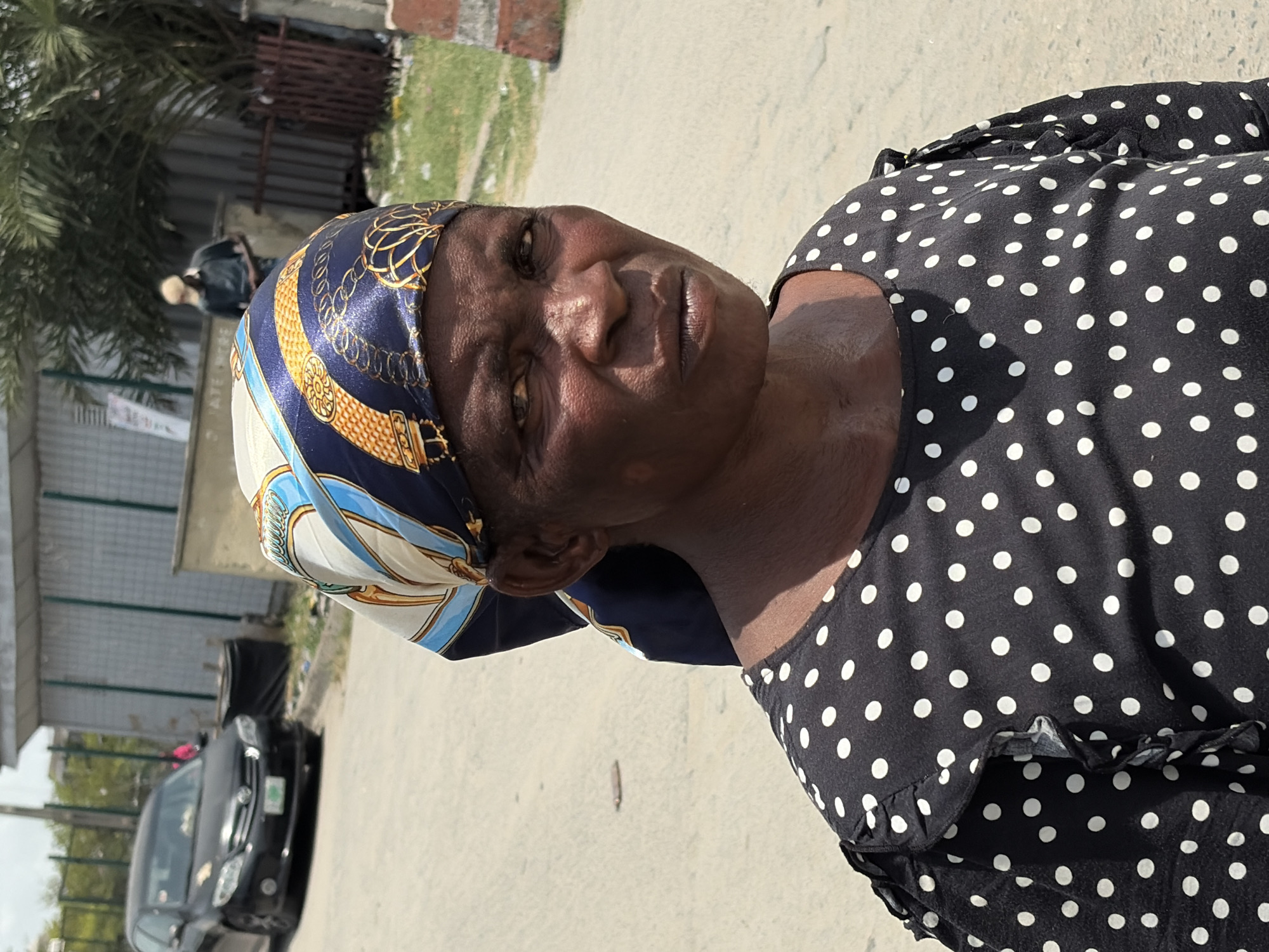 A woman wearing a navy blue and white patterned headscarf and a black and white polka dot dress stands on a concrete surface in Igboefon, Nigeria. A black car and fence are visible in the background.
