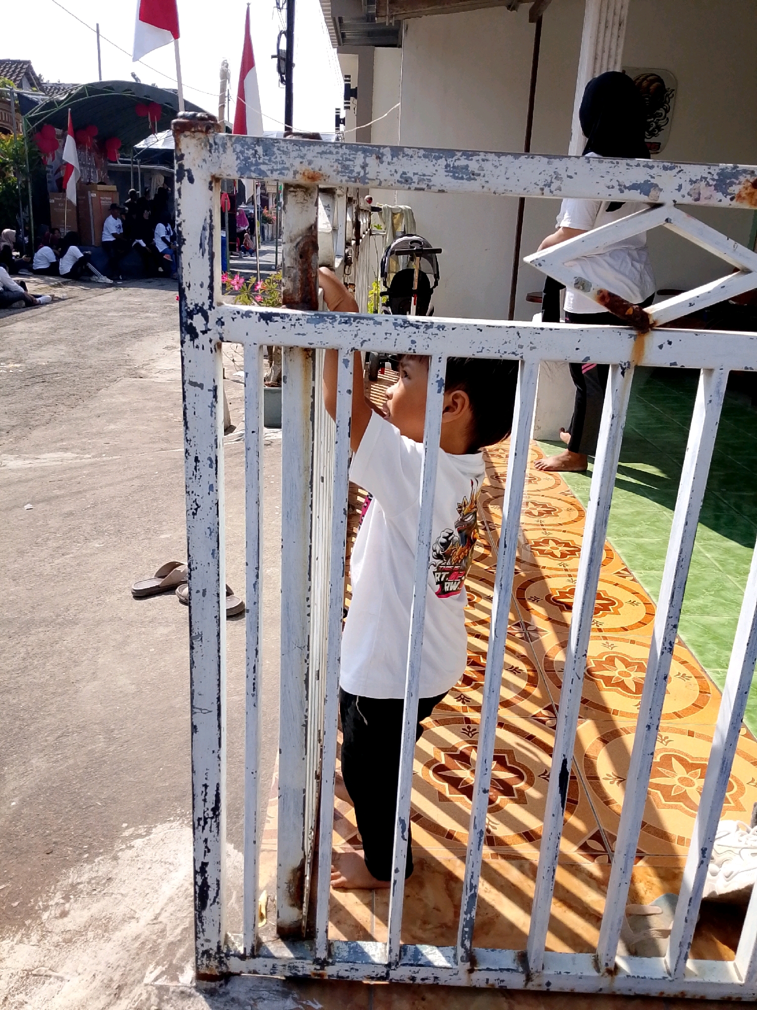 In Banyakan, Indonesia, a young child wearing a white t-shirt and black pants stands behind a white metal gate, reaching out to touch the gate. The child's face is turned away from the camera, and they are positioned to the right of the gate's opening. The background reveals a paved area with some foliage and a building with a tiled floor. Several people are visible in the distance, seated on the ground, and Indonesian flags are displayed. The lighting suggests it is daytime, with strong sunlight creating shadows.