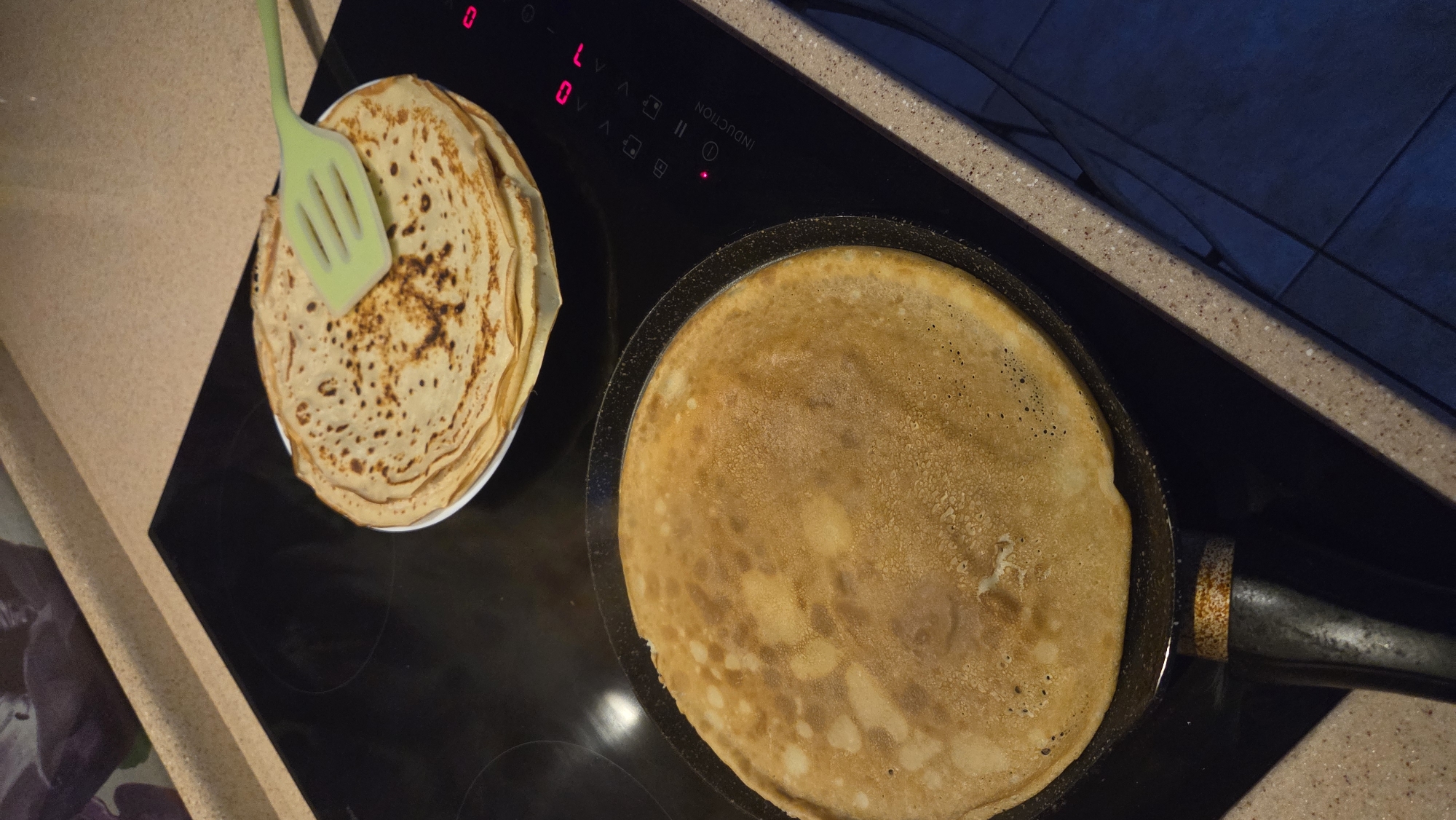 An overhead shot captures a domestic scene in a kitchen in Chaikovsky, Russia, likely during the evening or night due to the artificial lighting and shadowed background. The primary subjects are food and cooking equipment.
On the left, a stack of golden-brown pancakes or crepes is neatly piled on a white plate, indicating a batch has already been prepared. A light green silicone spatula rests on top of this stack. To the right, a single large pancake is actively cooking in a black frying pan, its surface showing light browning and small air bubbles.
Both the plate of pancakes and the frying pan are positioned on a sleek, black induction stovetop. The stovetop features a touch control panel with illuminated red digital displays and icons. Visible text on the stovetop includes "INDUCTION" written vertically, "L" and "0" as power level indicators, and various universal icons for functions like power, pause, and timer. A small red indicator light is also on. The stovetop is set into a speckled beige and brown countertop. The background behind the stovetop shows glimpses of dark blue or grey tiled walls. The overall impression is one of active home cooking.