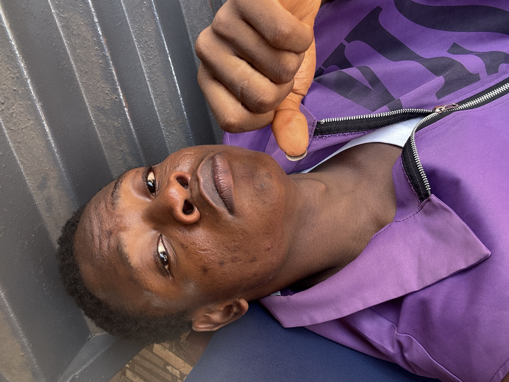 A dark-skinned person, with short black hair and visible pores, is lying on what appears to be a blue surface. The person is wearing a purple jacket, partially zipped, revealing a white undershirt. An arm and hand are held above the person's face. In the background are grey, striated metal panels. The location is Unknown, Unknown.