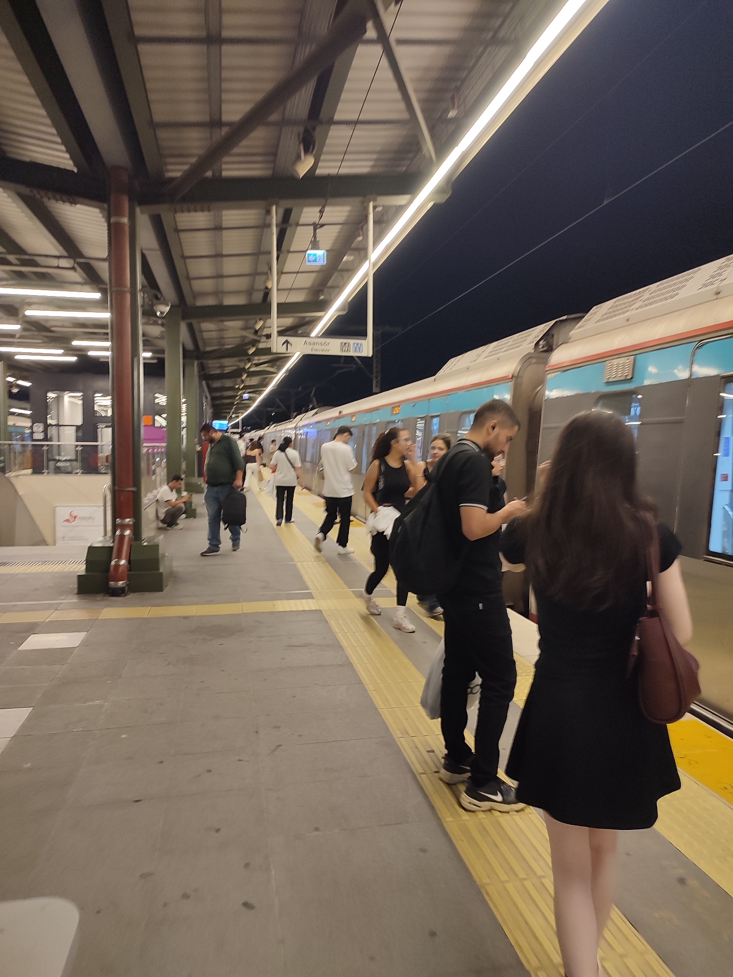 A group of people are standing on a train platform at night. A train is visible next to the platform. The platform is covered by a roof supported by columns. There are lights on the platform and under the roof. Signs indicating "Asansör" (Elevator) and an arrow pointing up are visible. Some people are holding bags and one person is looking at their phone. The floor of the platform has tactile paving.