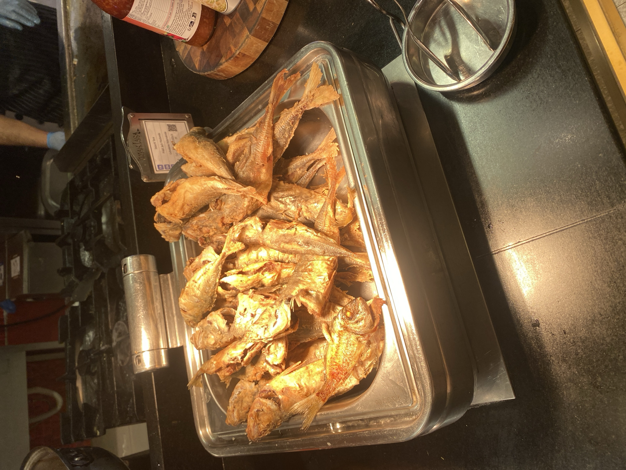 A metal serving tray filled with fried fish is displayed on a black countertop. The fish appear to be whole and golden brown. To the right of the tray, a small metal tongs rests in a shallow metal dish. Behind the serving tray, a sign is partially visible with text in Turkish and English, identifying the dish as "Fried Jack Mackerel". A bottle of red sauce is also visible to the right of the sign. The background shows a commercial kitchen with stainless steel appliances and equipment. A person wearing blue gloves and a striped apron is visible in the upper right corner of the image, suggesting they are working in the kitchen. There is no information in the image that suggests a specific location in the city of Aksu, Turkey.