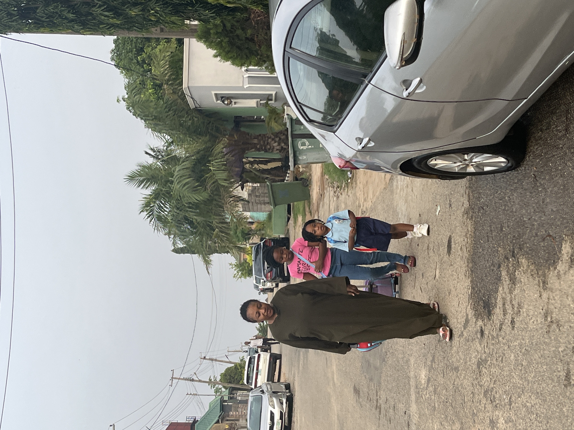 An outdoor, daytime photograph captures a street scene in Abuja, Nigeria, under an overcast sky. In the foreground to the left, a silver car is parked on a paved street, with its rear side and passenger doors visible. To the right of the car, four individuals are standing or walking along the side of the road.
The prominent figure is a woman in the midground, wearing a long, dark green or brown robe-like garment. She is facing forward, looking towards the viewer with a neutral expression. Behind her and slightly to her right are three young girls. One girl, in a blue and white school uniform, stands furthest back. Another girl in front of her wears a bright pink t-shirt with the text "MAMA GIRL" printed on it, paired with blue jeans. The third girl, mostly obscured by the woman, also appears to be wearing casual attire.
The background features residential buildings with light-colored facades and green roofs, interspersed with dense green trees and palm trees. Utility poles with multiple power lines are visible against the pale sky. Further down the street, more parked cars and distant figures can be seen, indicating an active neighborhood. The overall impression is a typical street in a residential area, possibly during a casual outing or a school run. The weather is bright but overcast, with no direct sunlight.
