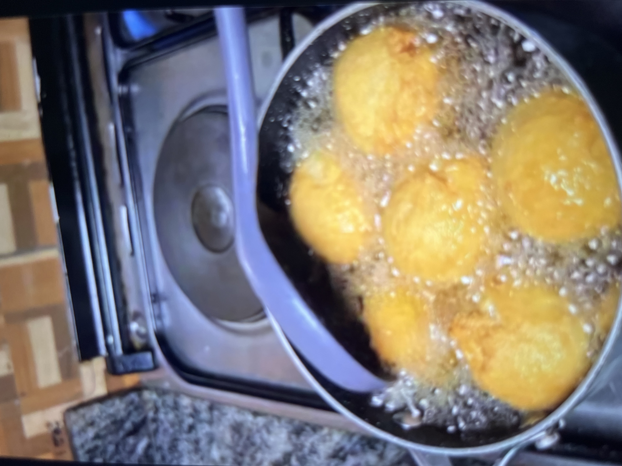In Abuja, Nigeria, a close-up shot captures a pot of golden-brown fritters being deep-fried in bubbling oil on a stovetop. The fritters, several of which are visible and submerged in the hot oil, have a rounded, slightly irregular shape. A pale purple ladle rests on the edge of the pot, its tip submerged in the oil, ready to stir or remove the fritters. The stovetop appears to be a standard electric model with black circular heating elements. The background shows a tiled kitchen wall with a brown and beige pattern, and a hint of a granite-patterned countertop to the left. The lighting suggests the image was taken indoors, possibly during the daytime, given the overall brightness.