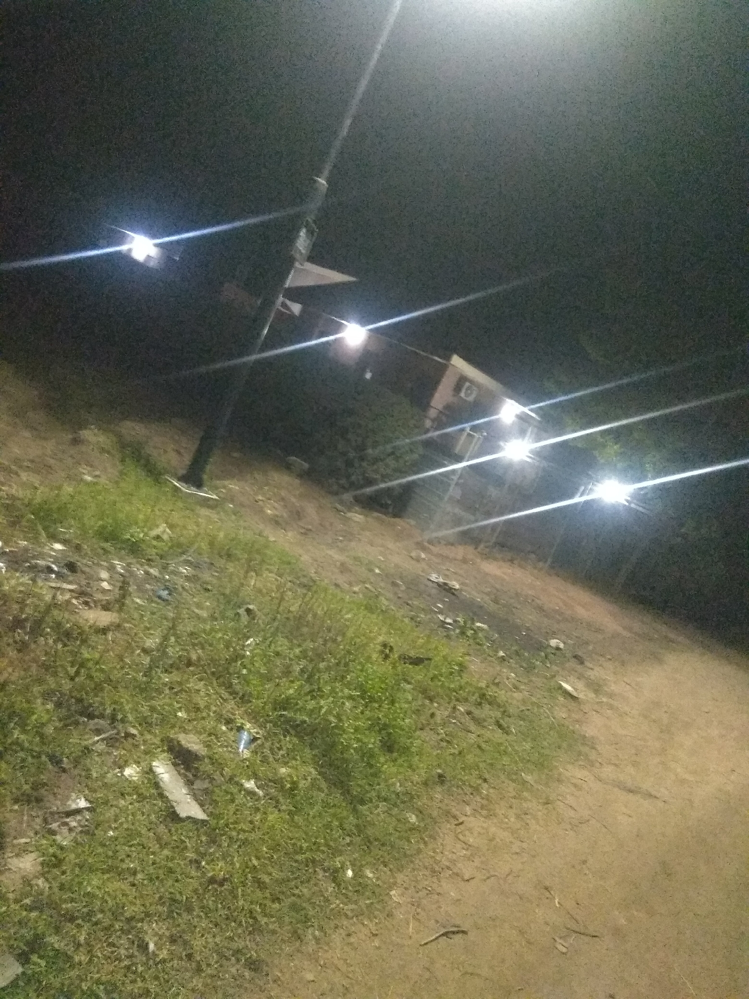 The image depicts a nighttime scene in Jalingo, Nigeria, likely a roadside or undeveloped area. A prominent street lamp with multiple lights illuminates the foreground and midground, casting harsh light beams that streak across the frame. The ground is uneven, with patches of green grass and dirt. Litter is scattered on the ground, including what appears to be plastic and debris. In the midground, a small building with lights visible is situated among trees. The overall impression is one of a utilitarian, sparsely populated outdoor area illuminated by artificial light.