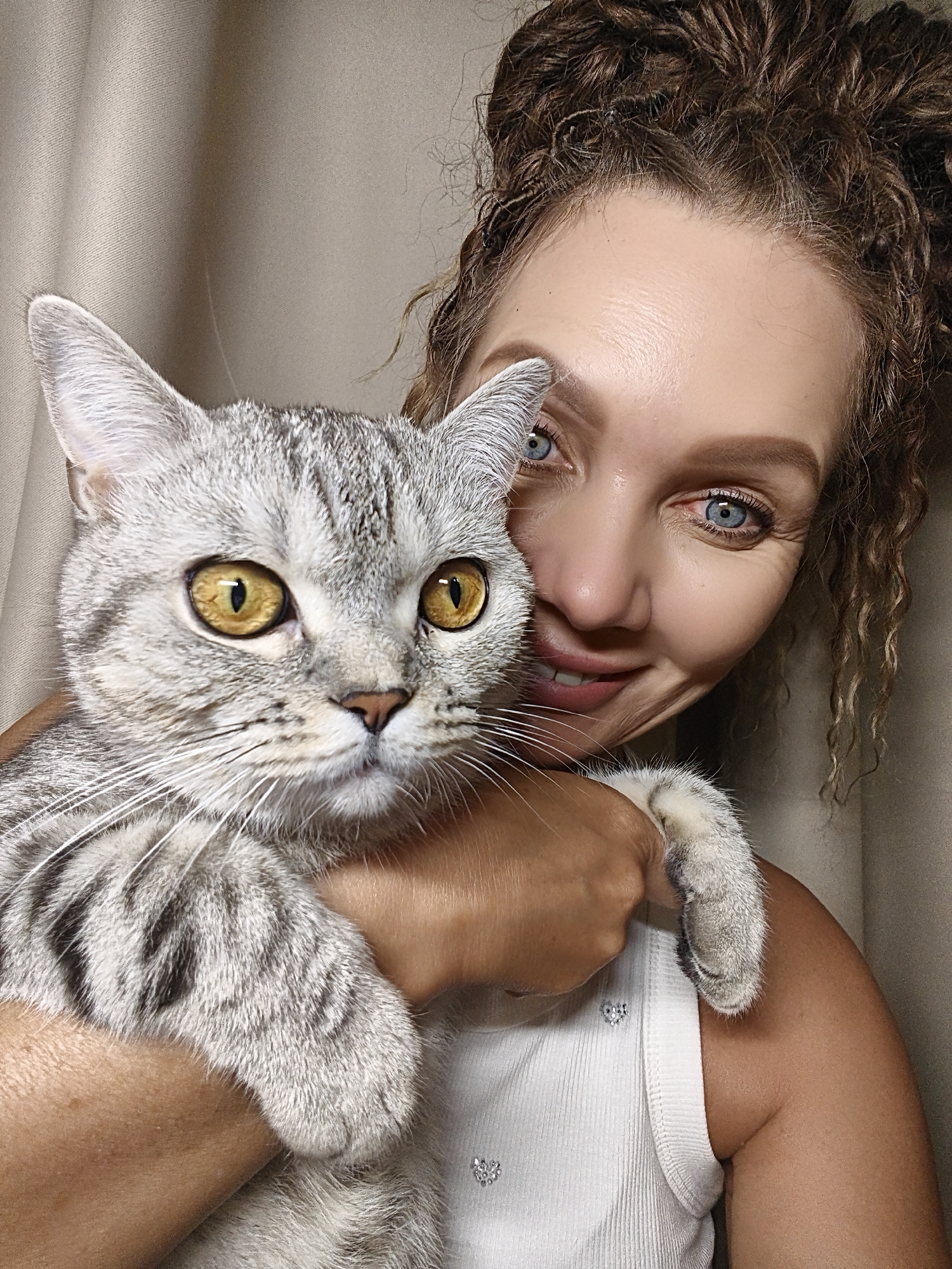 In an indoor setting with a neutral-toned curtain in the background, a woman with curly brown hair styled in a bun and piercing blue eyes is holding a silver tabby cat. The woman is smiling and looking towards the camera, with her head tilted slightly. The cat, with striking yellow eyes, is positioned in front of her, partially obscuring her face. The woman is wearing a white ribbed tank top adorned with small, sparkling heart-shaped embellishments. The cat's paw is visible resting on the woman's shoulder. The image is a close-up shot, focusing on the interaction between the woman and her pet.