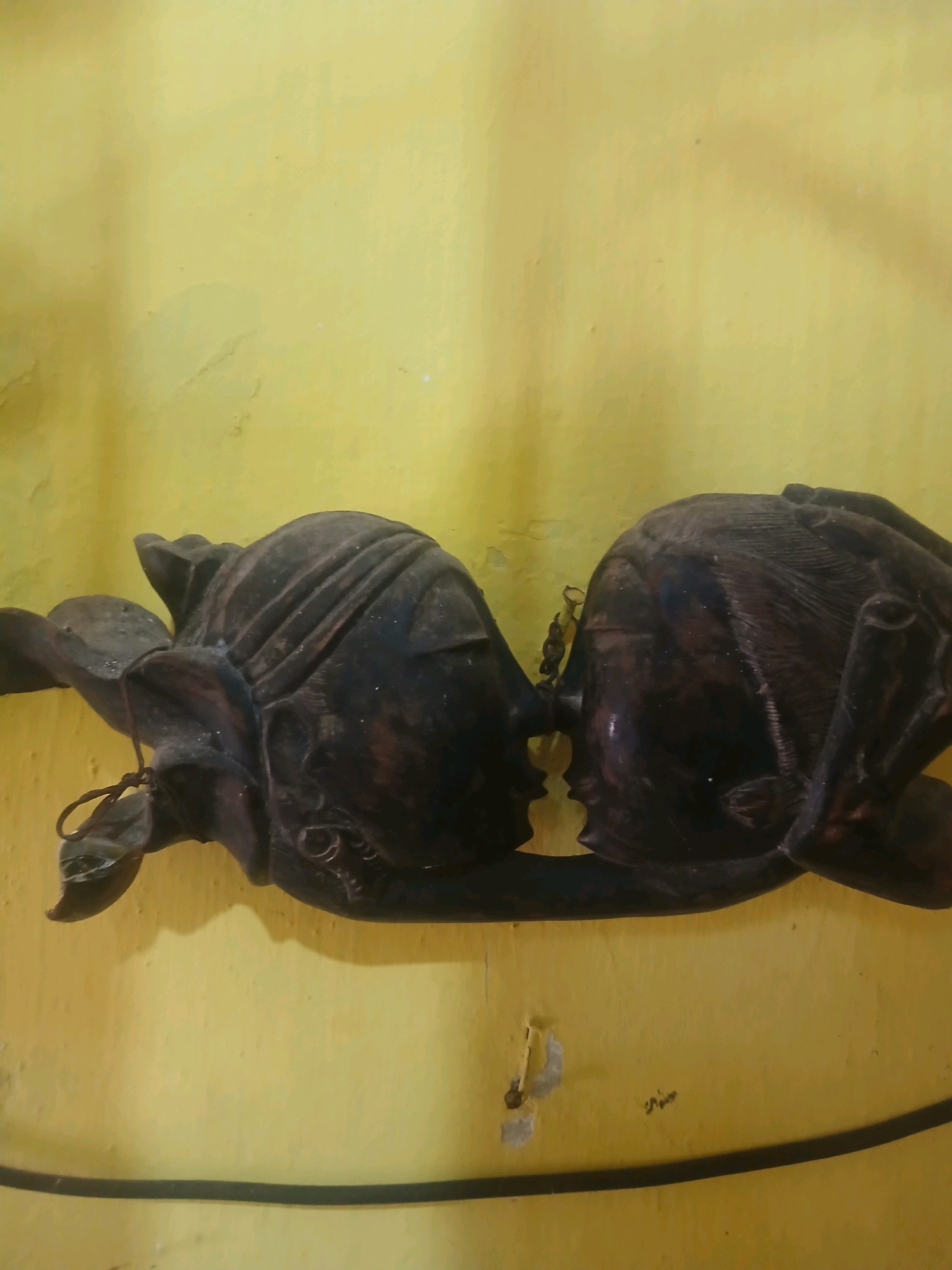 A carved wooden sculpture of two faces, possibly representing figures from Indonesian folklore, is mounted on a textured yellow wall. The faces are dark brown and appear to be in profile, leaning towards each other as if to kiss. The left face has a leaf-like adornment attached by a string, while the right face has intricate carvings on its head. The sculpture is mounted horizontally. The yellow wall shows some signs of wear and tear, with patches of lighter color and a small nail visible in the lower section. A thin black line, possibly a border or part of a frame, runs horizontally across the lower portion of the image. The lighting is somewhat dim, casting shadows on the wall and highlighting the details of the carving. This scene is located in Banyuwangi, Indonesia.
