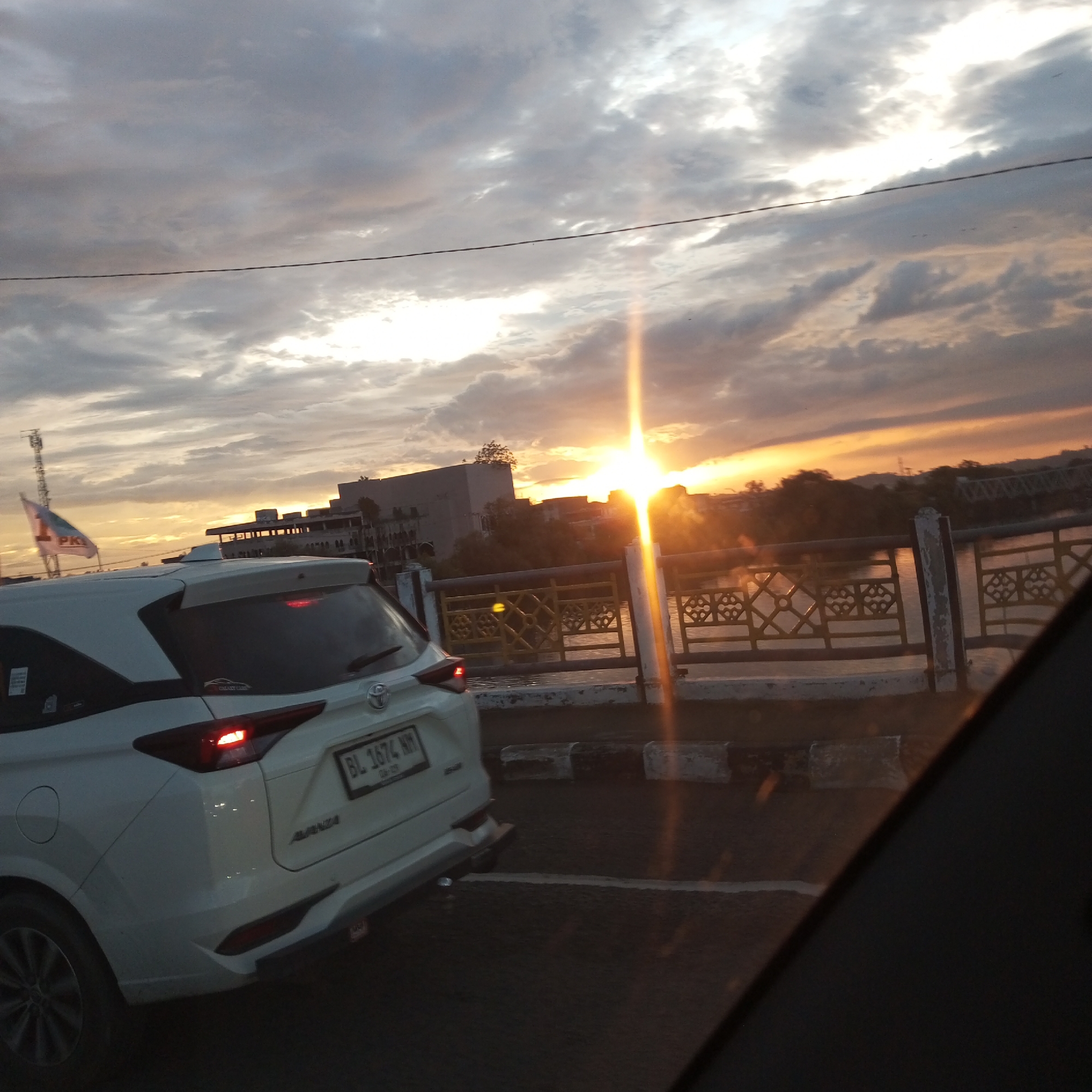 The image shows a white Toyota Avanza car on a bridge in Lhokseumawe, Indonesia at sunset. The car has a license plate number BL 1674 NW. The bridge has a decorative yellow and white railing and overlooks a body of water. Buildings are visible in the background, along with a flag. The sky is cloudy and the sun is setting, creating a bright glare. The image appears to be taken from inside another vehicle, as part of a dark dashboard is visible in the foreground.
