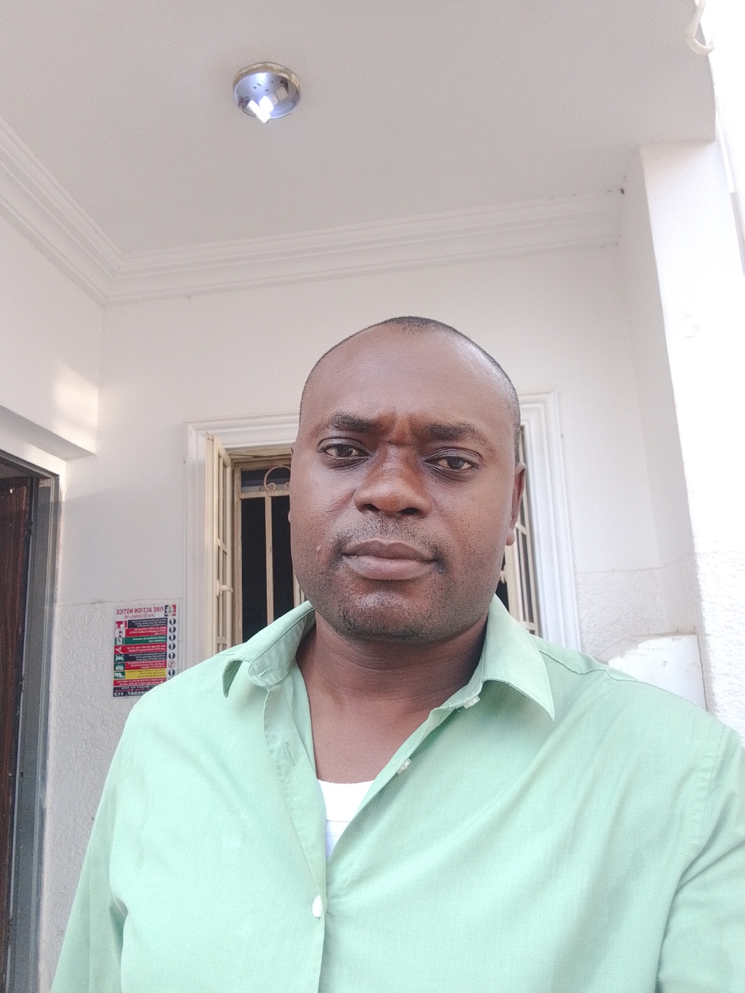 A medium shot of a Black male wearing a light green, collared button-down shirt, standing outdoors against a white building. The background includes a white wall with a framed window and a door to the left. A small sign is affixed to the wall to the left of the window. The location is in Gwarinpa, Nigeria.