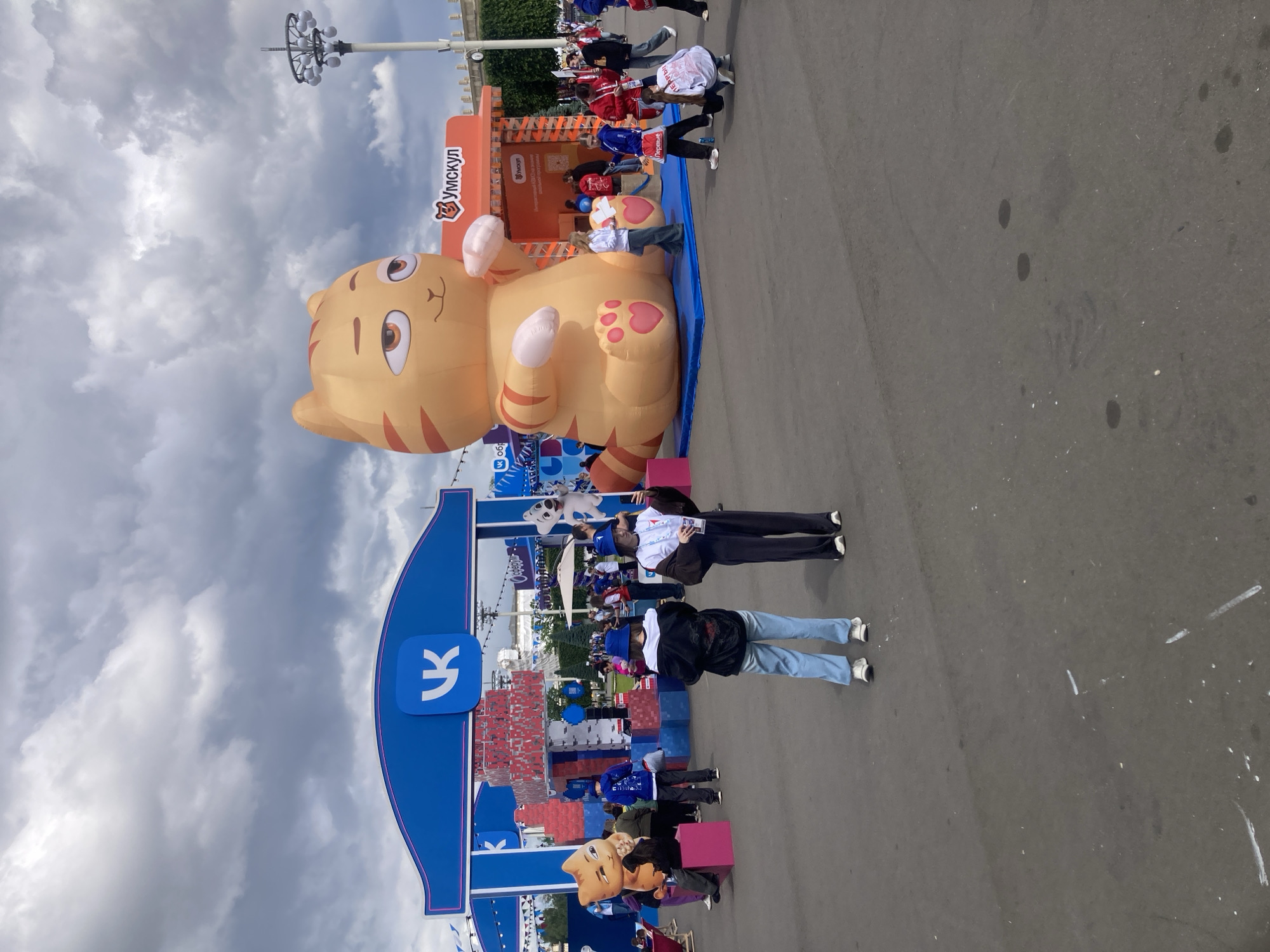 This photograph shows a public event in Moscow, Russia. A large, orange inflatable cat figure, labeled "Умскул," is the focal point. People are interacting with and posing near the inflatable. A blue structure with a stylized "K" logo is also prominent, potentially indicating a sponsor or brand. The background includes a crowd of people, other promotional materials, and overcast skies. The ground appears to be pavement or asphalt.